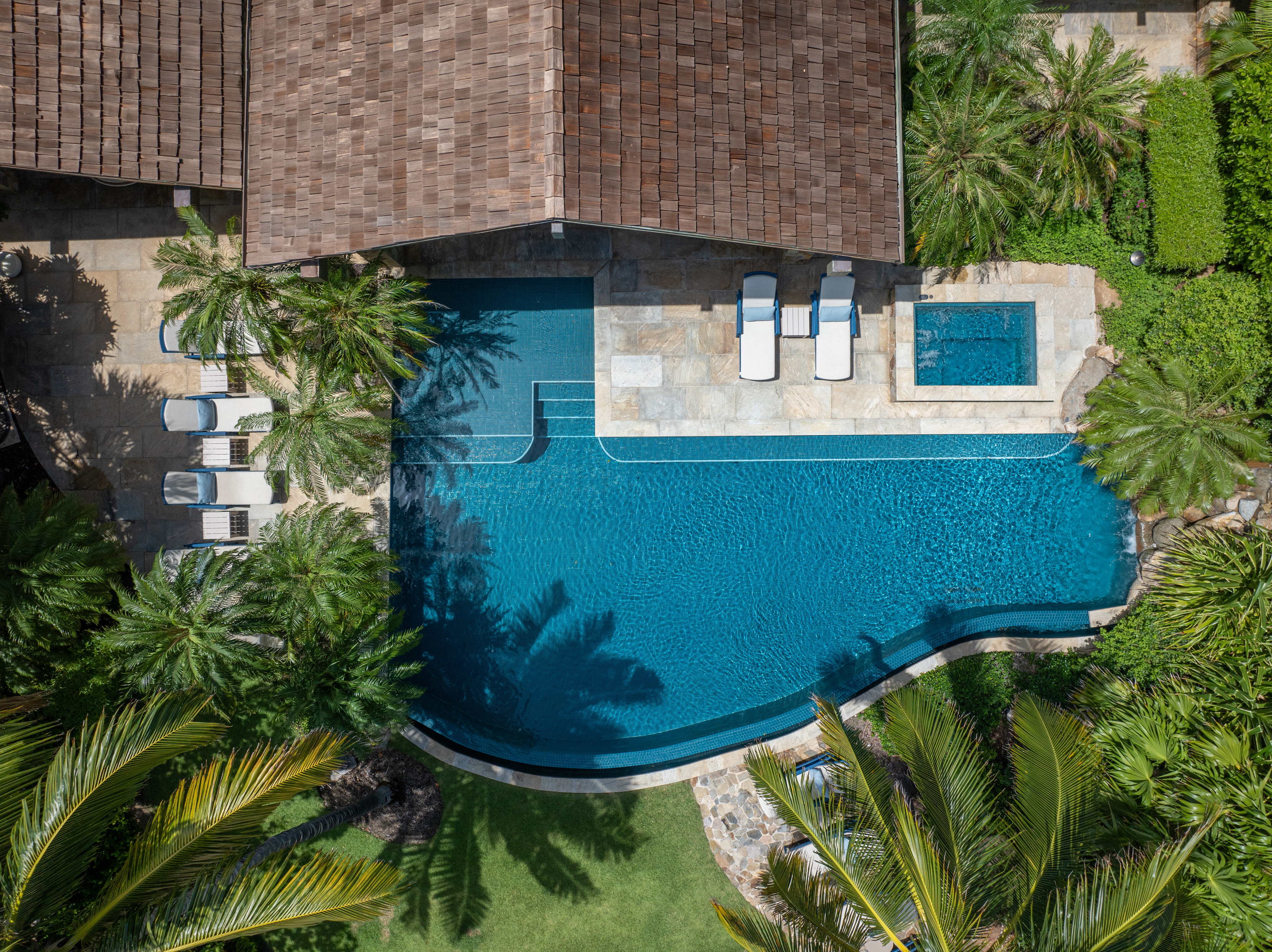 Top-down view of Nonamé Villa's backyard with a large, irregularly shaped swimming pool. The pool area features a hot tub, lounge chairs, and is surrounded by lush greenery and a paved deck. A portion of the house with its tiled roof borders the inviting pool area.