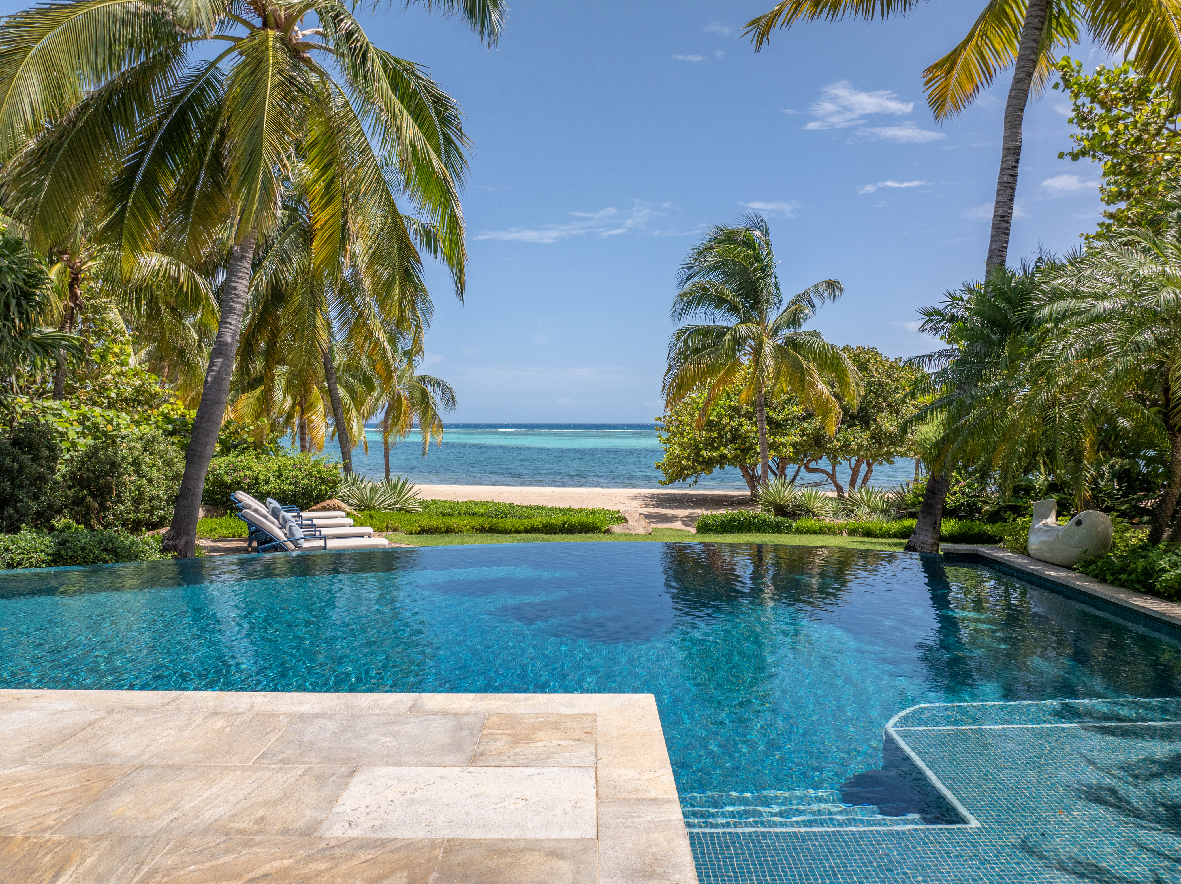 A luxurious infinity pool at Nonamé Villa overlooks a serene beach with white sands and clear blue waters. Palm trees and lush greenery surround the pool area. Lounge chairs and a beachside path lead toward the ocean under a bright, sunny sky.