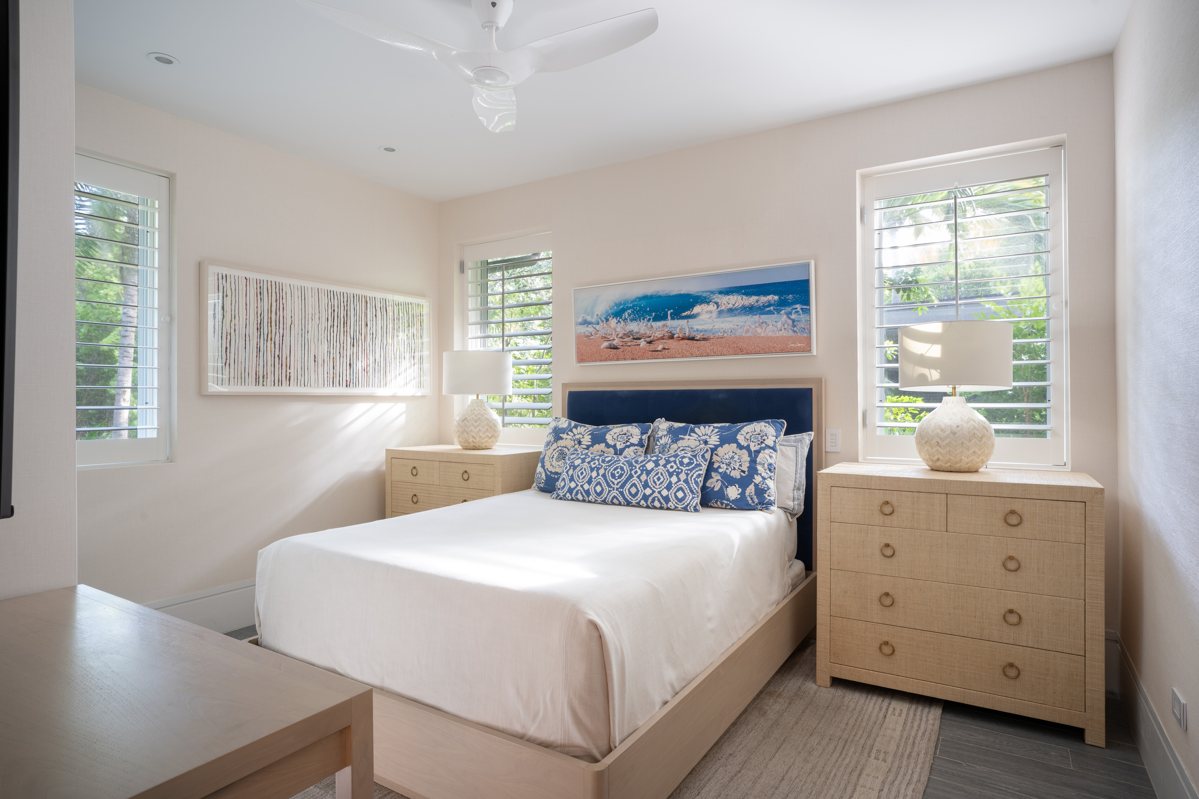 A bright bedroom in Nonamé Villa features a queen-sized bed centered between two wooden nightstands, each with a spherical lamp. The bed is adorned with white linens and blue, patterned pillows. Two windows with plantation shutters light the room, which is decorated with beach-themed wall art.