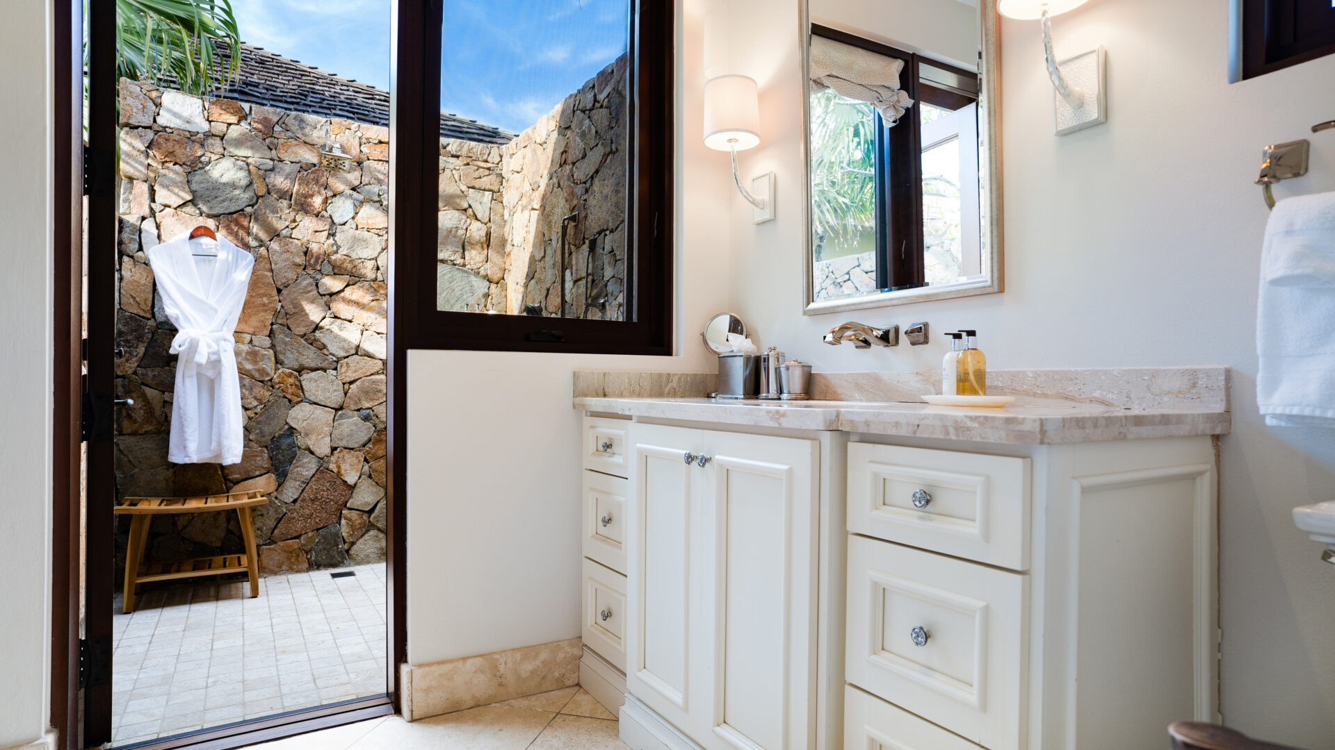 Spacious bathroom at Bella Beach Villa featuring white cabinetry and marble countertop, equipped with a sink and toiletries. A large window reveals an outdoor stone wall, while a doorway leads to an outdoor shower area where a robe hangs on a hook.