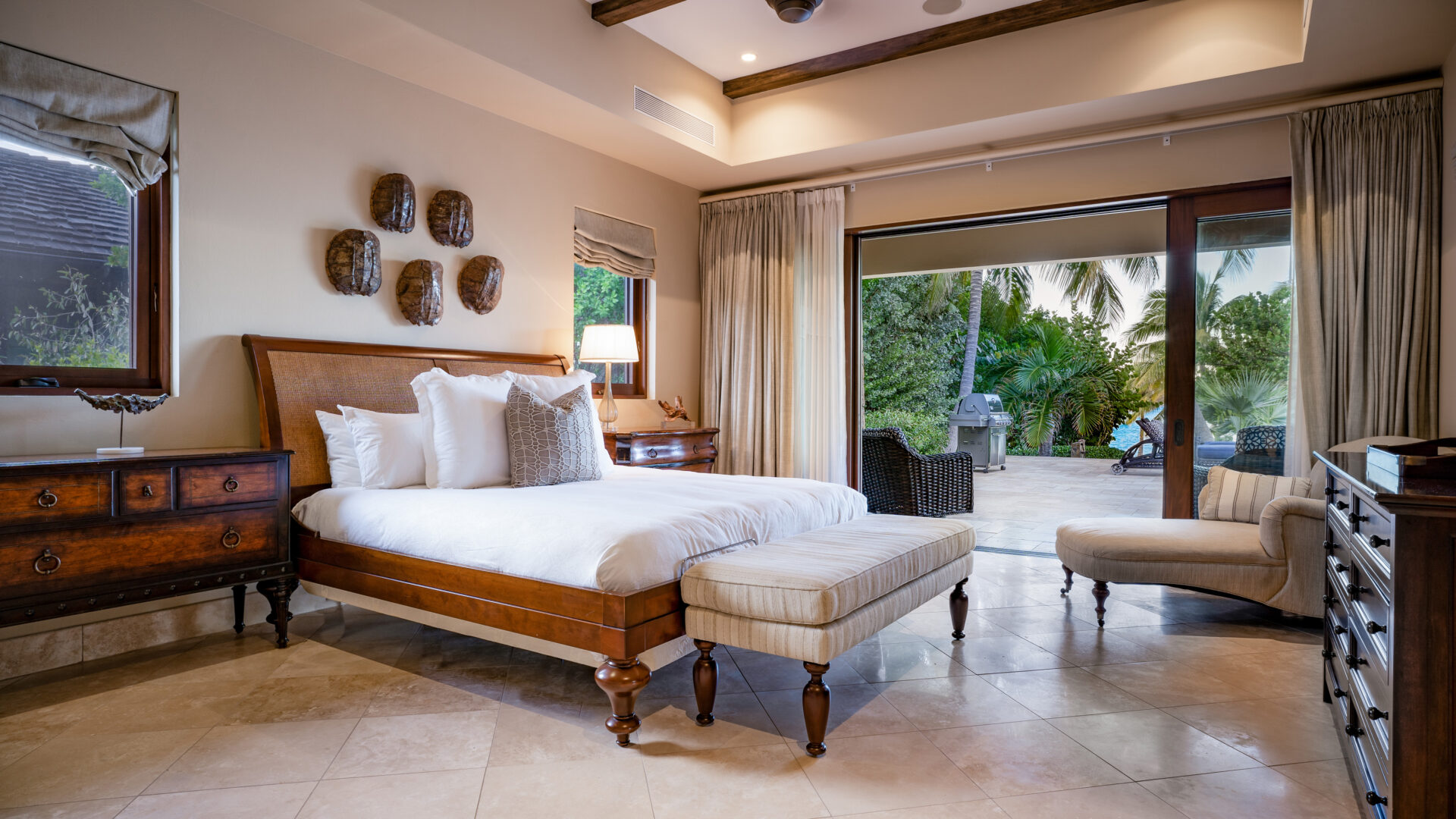 A luxurious bedroom at Bella Beach Villa with a large bed, wooden furniture, and beige tile flooring. The room features a view of a lush tropical garden visible through glass doors. Decorative wall masks, a cushioned bench at the bed's foot, and a cozy chair by the window complete the setting.