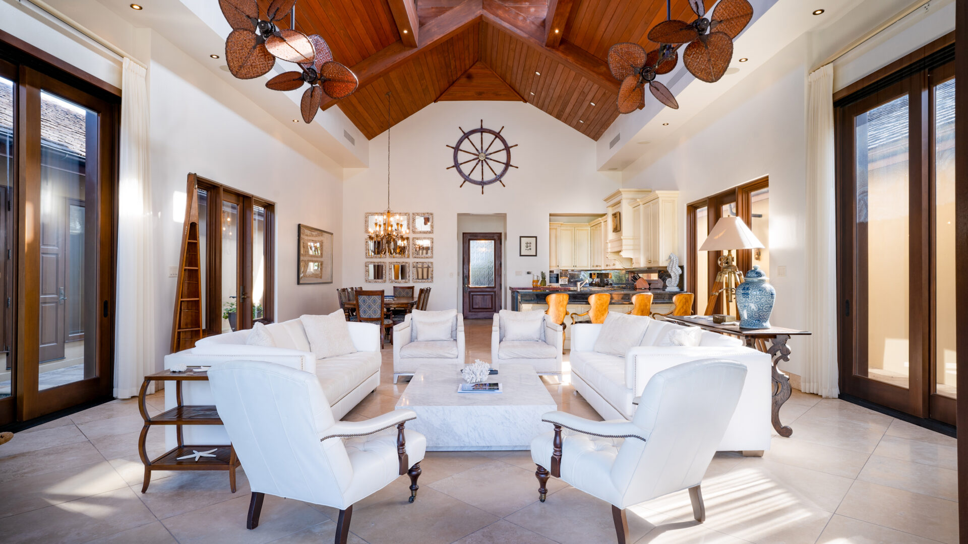 A spacious, well-lit living room at the Bella Beach Villa features a high wooden ceiling with three ceiling fans and large windows. White sofas and chairs are arranged around a central coffee table, under the ship's wheel decoration on the back wall, which faces the kitchen area.