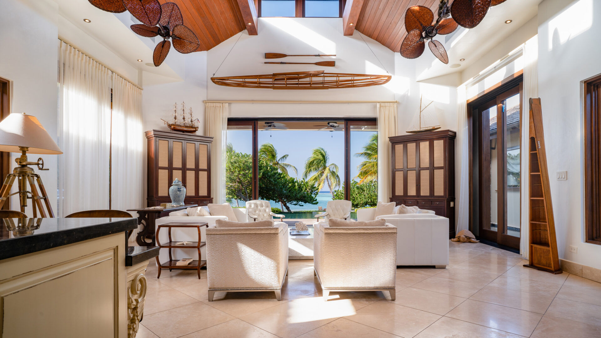 A spacious, well-lit living room with high wooden ceilings and large windows offering a view of Bella Beach Villa's tropical beach. The room features white furniture, wooden cabinets, ceiling fans, a wall-mounted wooden canoe, and a collection of nautical decor.