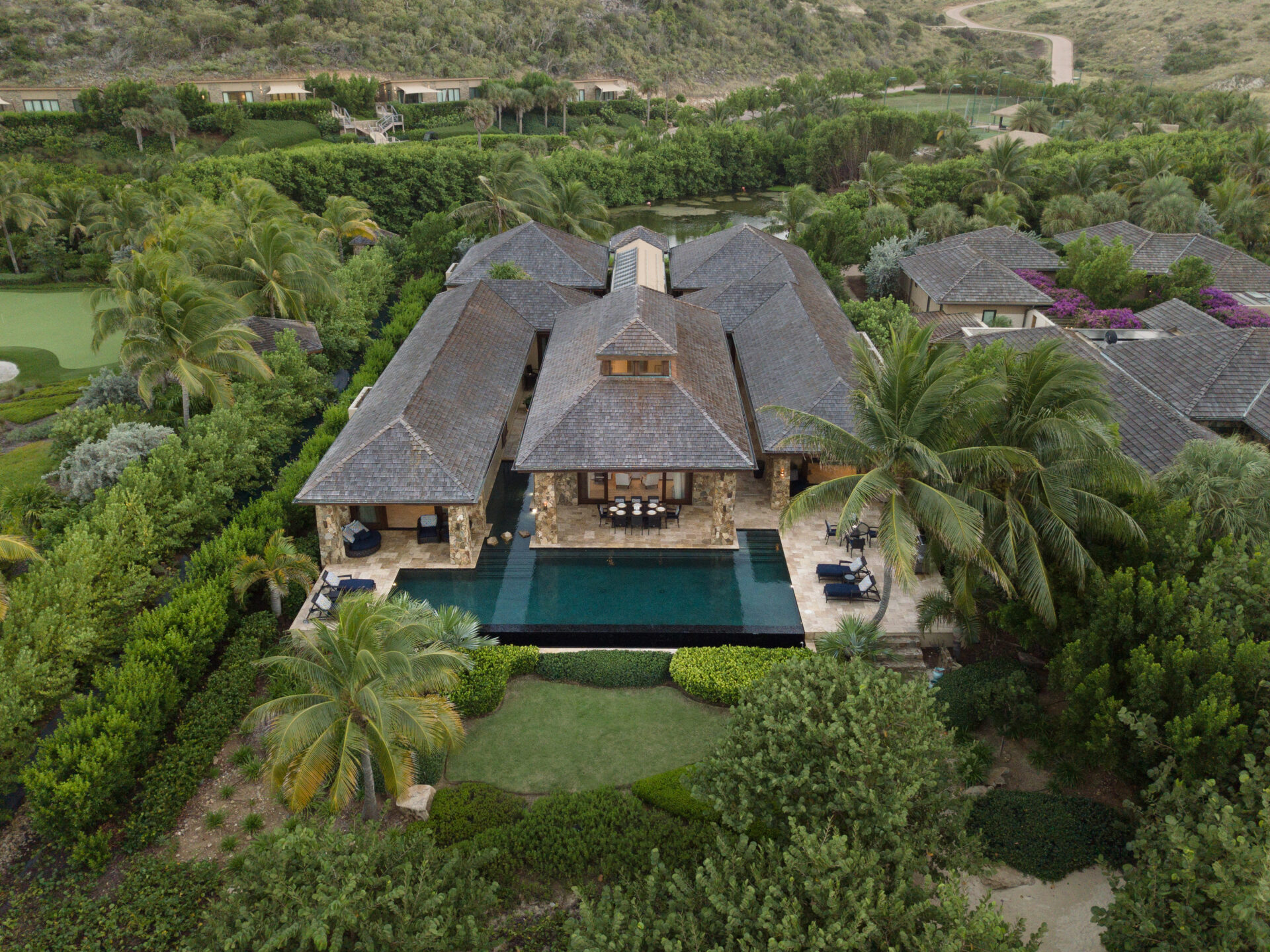 Aerial view of Bella Beach Villa, a luxurious mansion surrounded by lush greenery. The house has a dark tile roof and is centered around a large, rectangular pool. Palm trees and well-manicured shrubs enhance the landscape, with a golf course visible to the left.