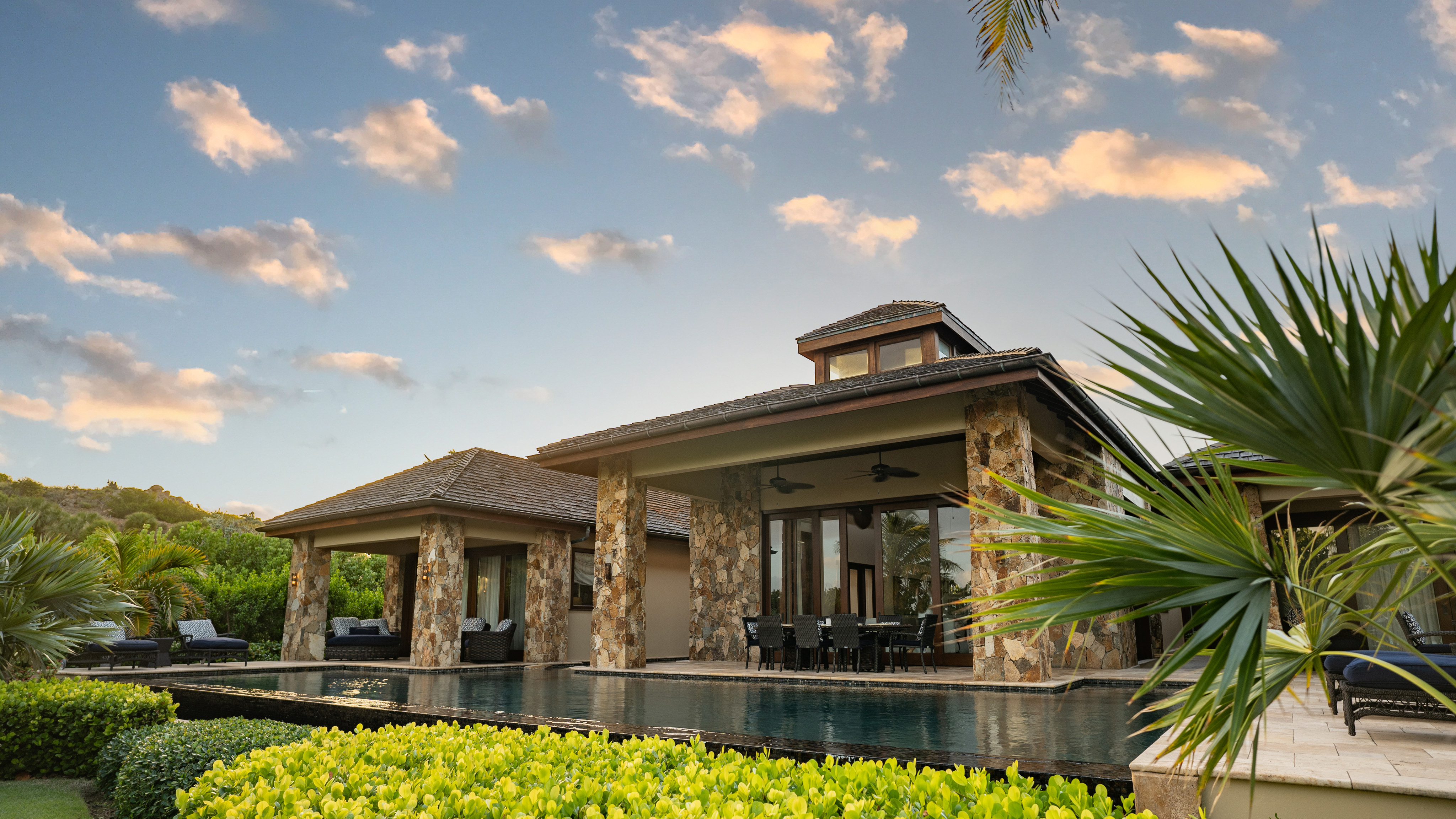 The Bella Beach Villa is a luxurious house with stone pillars, large glass windows, and a pitched roof set against a backdrop of a colorful sky with scattered clouds. It features an outdoor dining area, a pool, and lush greenery, creating a serene and elegant atmosphere.