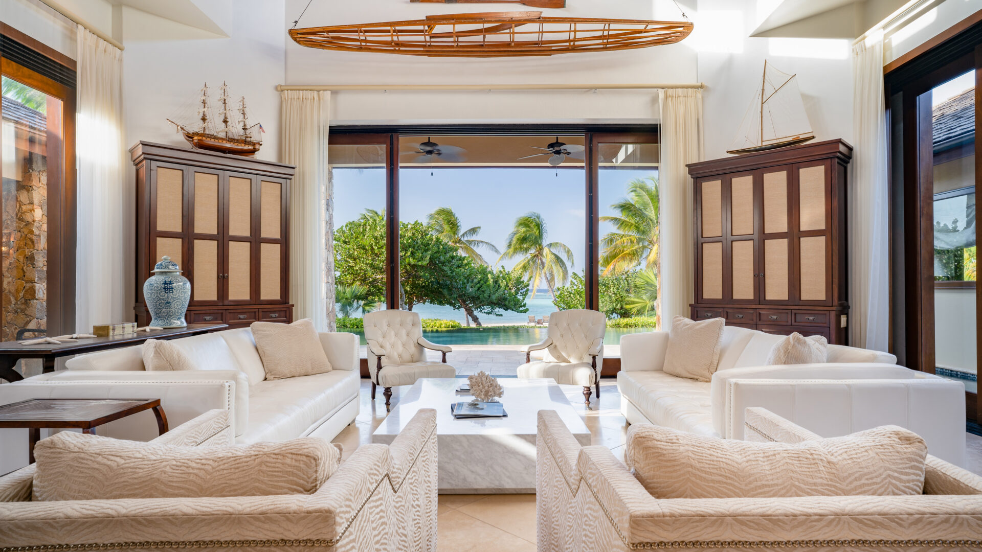 A bright living room with white sofas and armchairs, large windows revealing a tropical beach view with palm trees. The decor includes model sailboats, a decorative oar, and a blue vase. Sunlight streams in at the Bella Beach Villa, highlighting the airy and serene atmosphere.