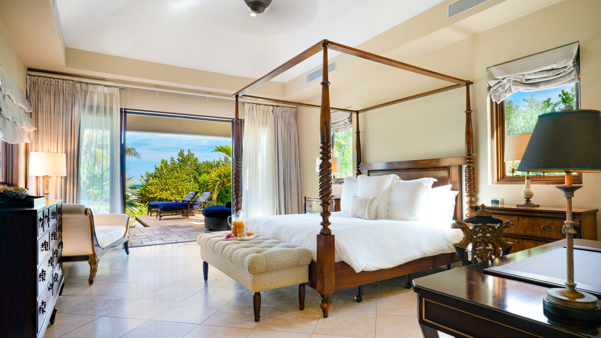 A luxurious bedroom at Bella Beach Villa features an elegant four-poster bed with white bedding, a cushioned bench at the foot, and wooden furniture. Large sliding doors open to a balcony with a lush garden view, allowing natural light to flood the room.