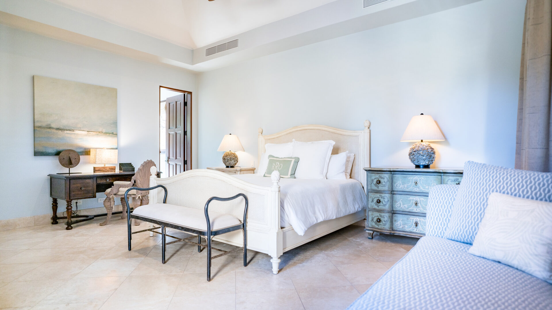 A bright, airy bedroom in Bella Beach Villa features a white wooden bed, light bedding, and two bedside tables topped with lamps. A bench sits at the foot of the bed. A cushioned chair is on the right side, and a painting hangs above a writing desk on the left. The room has a serene, calming atmosphere.