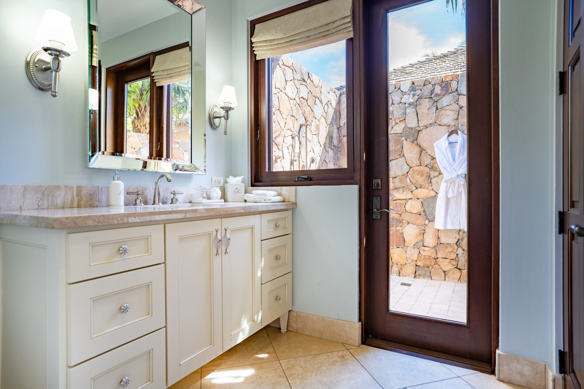 A bright, elegant bathroom at Bella Beach Villa features a large mirror, white cabinetry with a marble countertop, and modern wall sconces. A glass door opens to an outdoor stone wall, where a white robe hangs, and a pattern of light is cast onto the tiled floor.