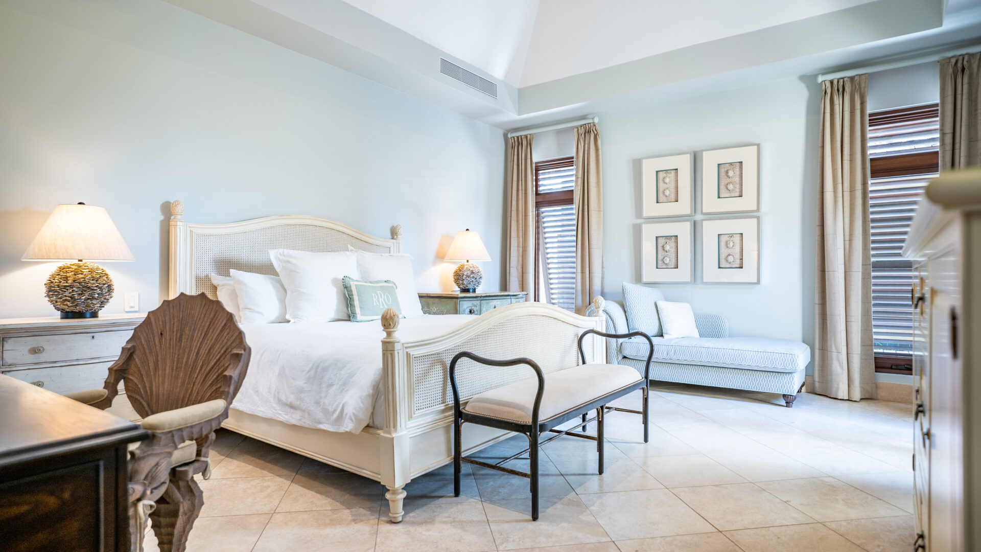 A bright and airy bedroom at Bella Beach Villa features a white bed with an upholstered headboard and footboard, bedside tables with lamps, a cushioned bench at the foot of the bed, framed art on the wall, and a cozy seating area by two large windows with beige curtains.