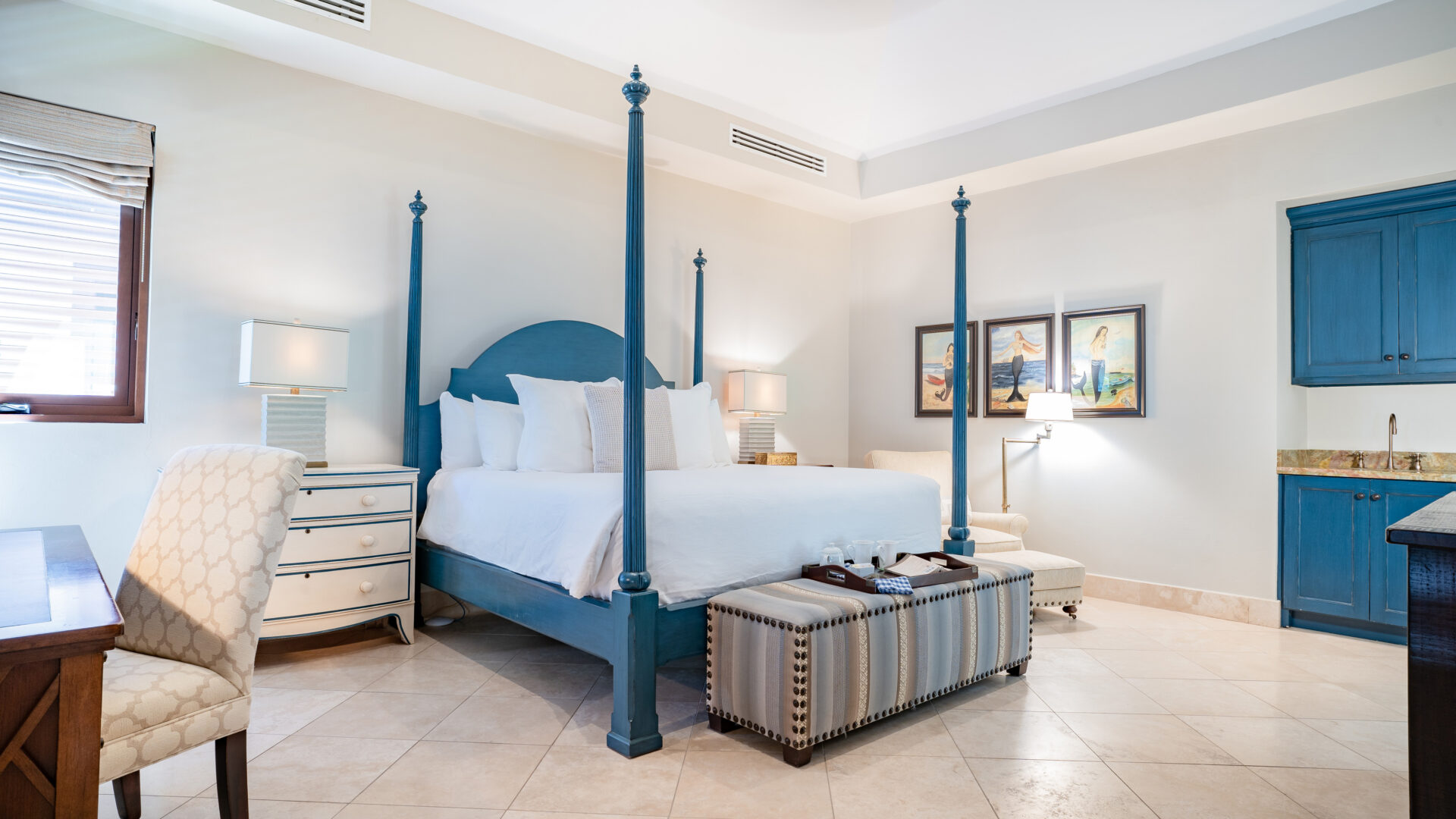 A spacious bedroom at Bella Beach Villa features a blue four-poster bed with white linens. To the sides of the bed are nightstands each with a lamp. The room also includes a striped ottoman, a desk, a beige armchair, framed wall art, and built-in blue cabinetry with a countertop.