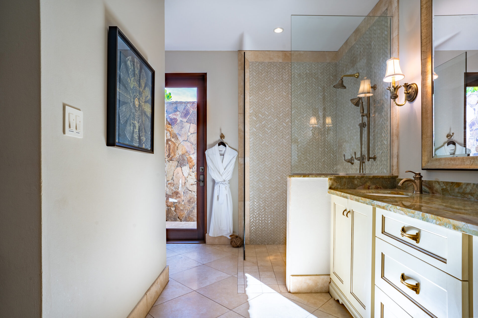 Luxurious bathroom in Bella Beach Villa with a walk-in shower featuring herringbone tile design. A white robe hangs by the wooden door leading outside. The vanity includes a marble countertop and under-mount sink with gold fixtures. Framed artwork decorates the cream-colored walls.