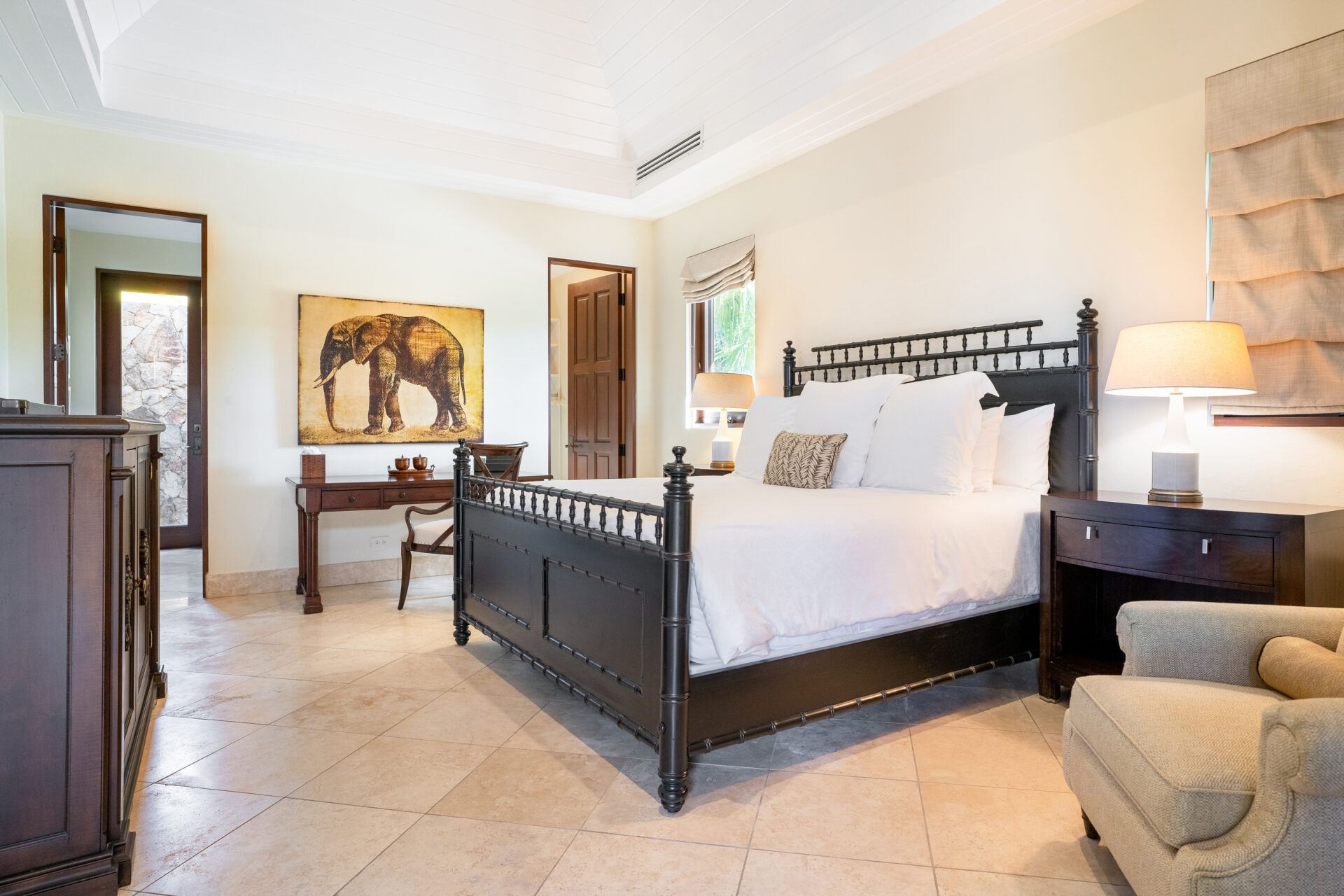 The Bella Beach Villa bedroom features a large black bedframe and white bedding. On the left, a wooden dresser and desk with a chair are complemented by an elephant painting above. The room boasts tiled floors, a beige armchair, nightstands with lamps, and light tan walls.