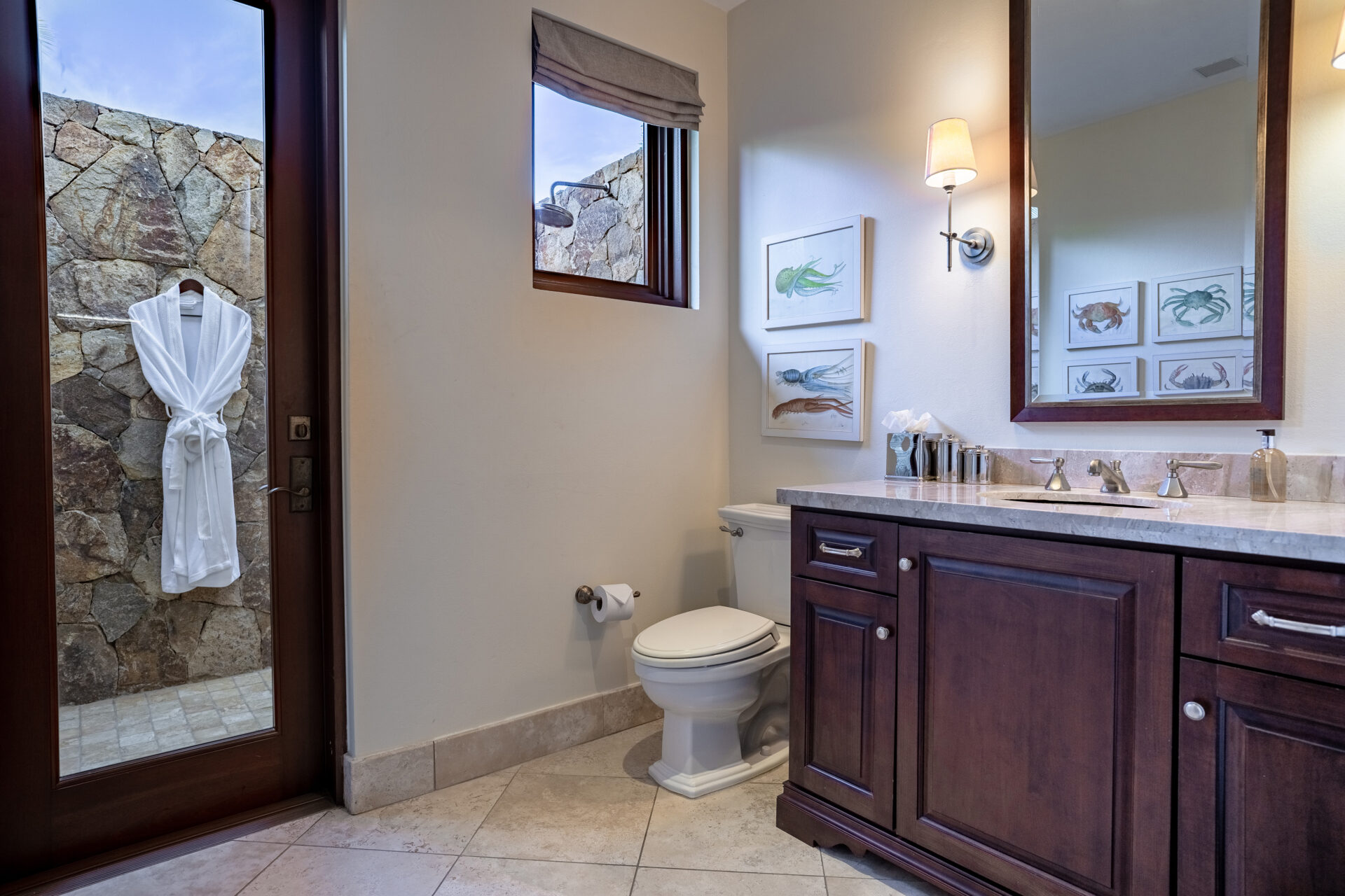 A modern bathroom in Bella Beach Villa features a wooden vanity with a marble countertop, a toilet, and framed sea life artwork on the wall. A door with frosted glass and stone walls leads to an outdoor shower area with a hanging white robe. Soft lighting and a small window add brightness.