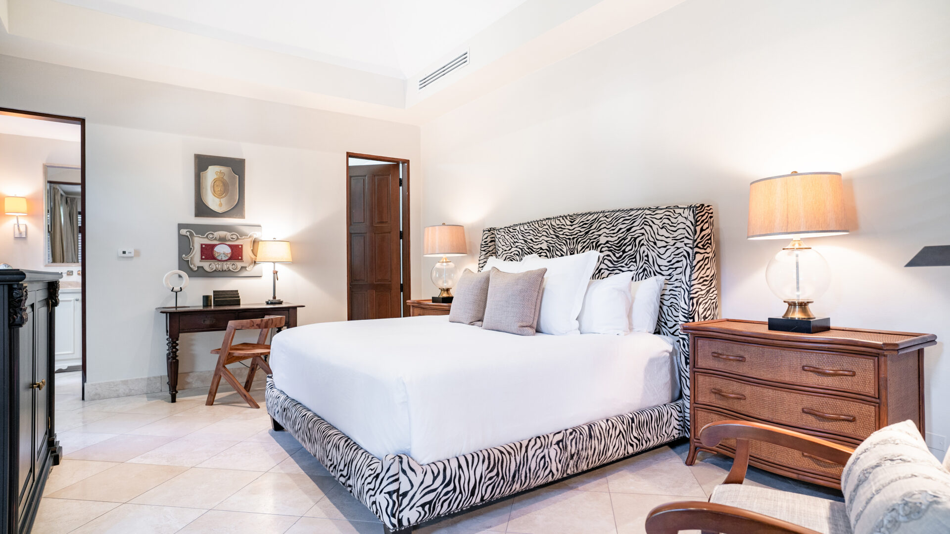 A modern bedroom at Bella Beach Villa features a large bed with a zebra-patterned headboard and white bedding. The room is decorated with wooden furniture, including bedside tables with lamps, a desk, and a dresser. A door to the bathroom is partially open, revealing a sink area.