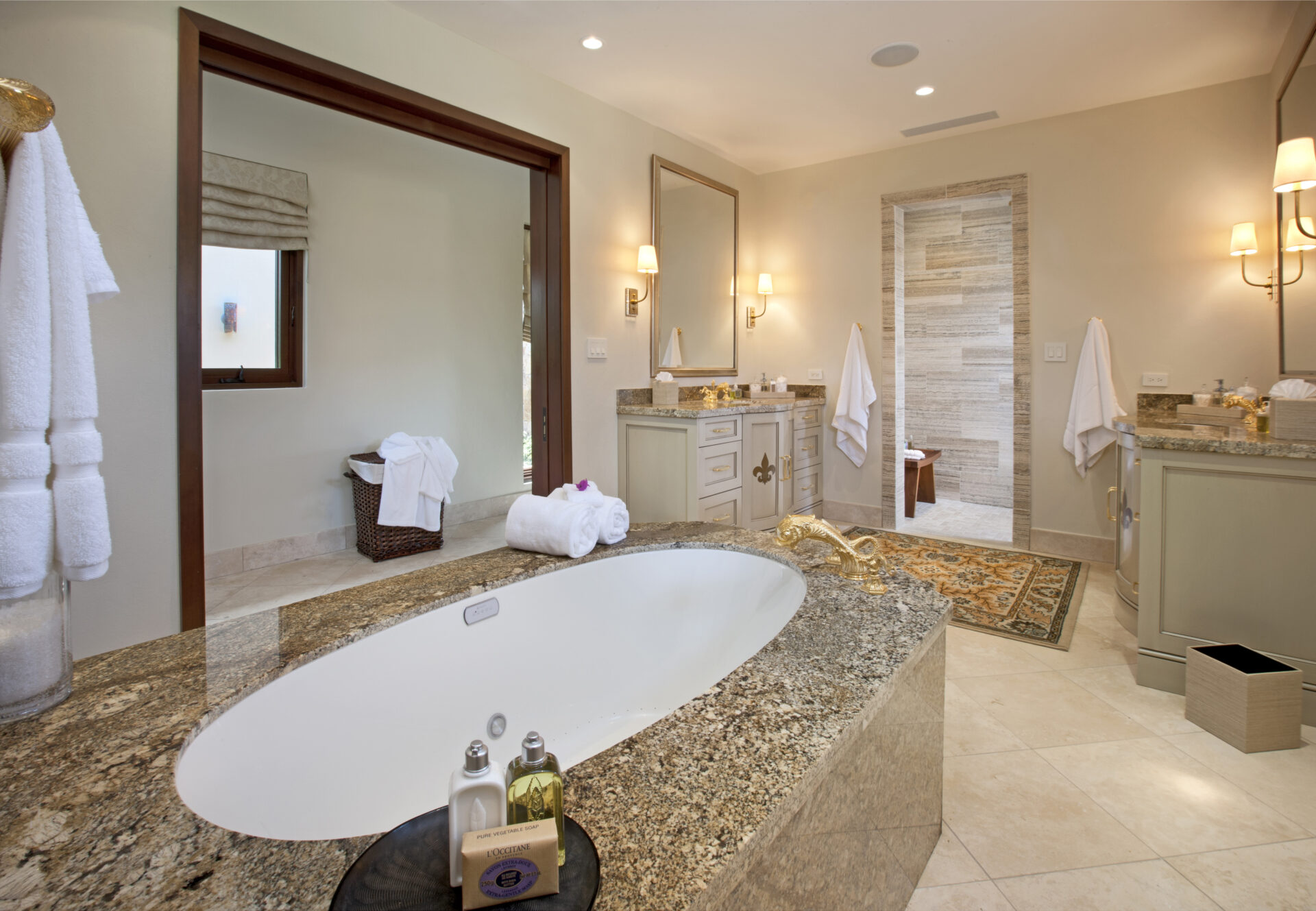 Luxurious bathroom at Bella Beach Villa with a large oval bathtub surrounded by granite countertops. The room features two separate vanities with illuminated mirrors, sconce lighting, and a separate shower area. Soft beige tones, white towels, and elegant decor create a serene ambiance.