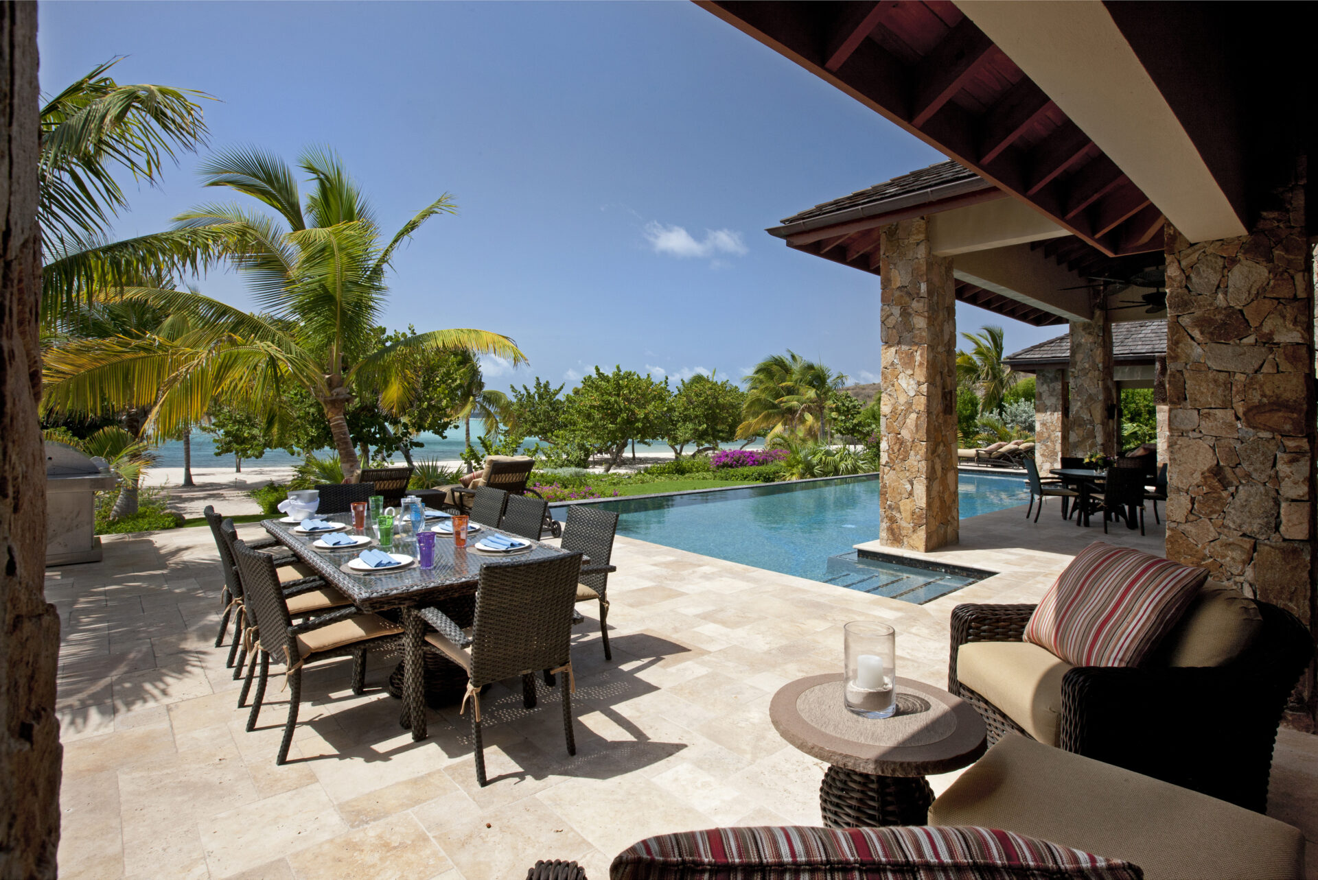 A luxurious outdoor patio at Bella Beach Villa features a dining table set for a meal, wicker chairs, and a comfy lounge area with striped cushions. The patio overlooks a serene swimming pool and a tropical garden with palm trees, leading to a sandy beach under a clear blue sky.