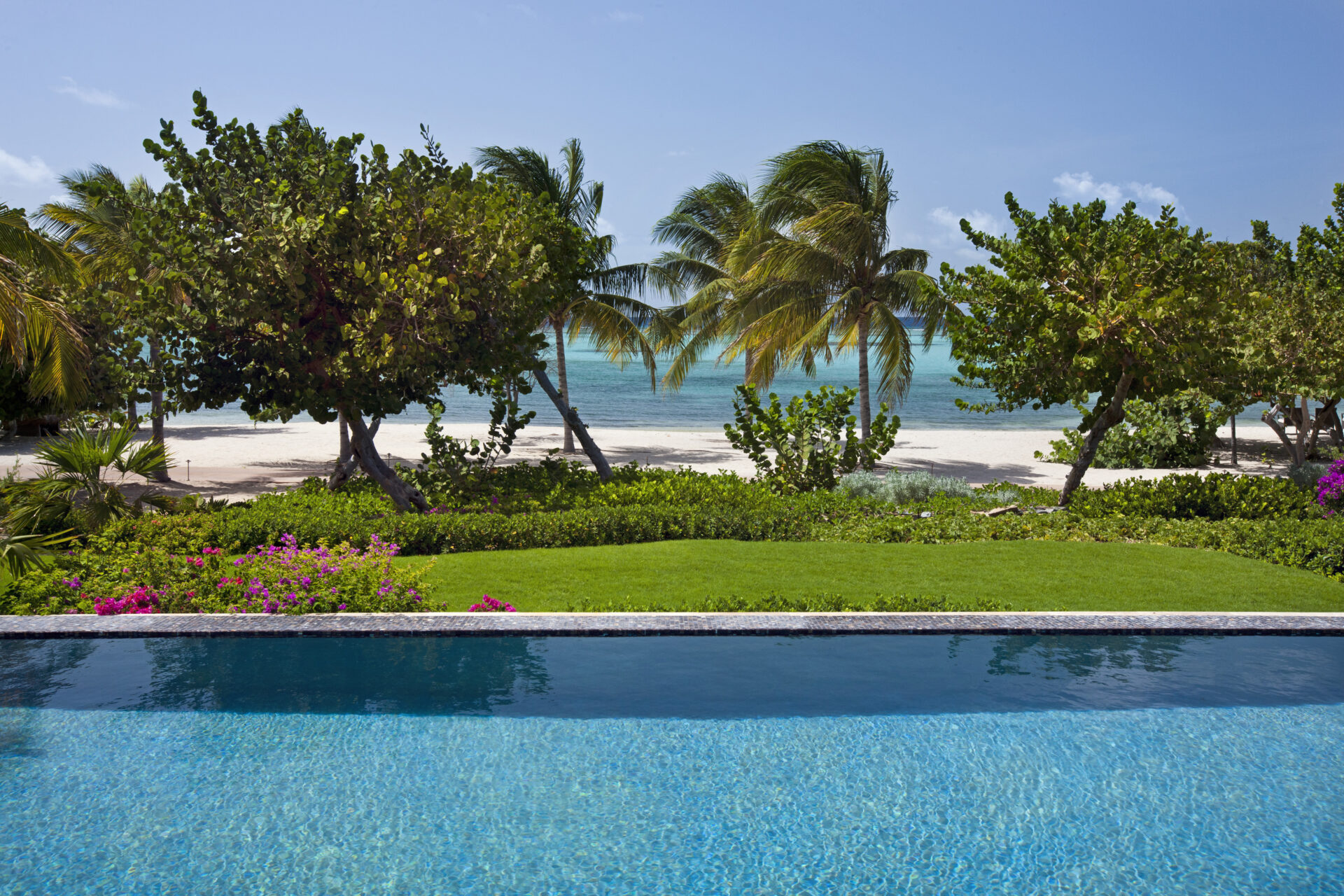 A tranquil seaside scene at Bella Beach Villa features a clear blue swimming pool in the foreground, lush green grass and vibrant flowering shrubs in the middle, and palm trees near a sandy beach by a calm turquoise ocean under a clear blue sky.