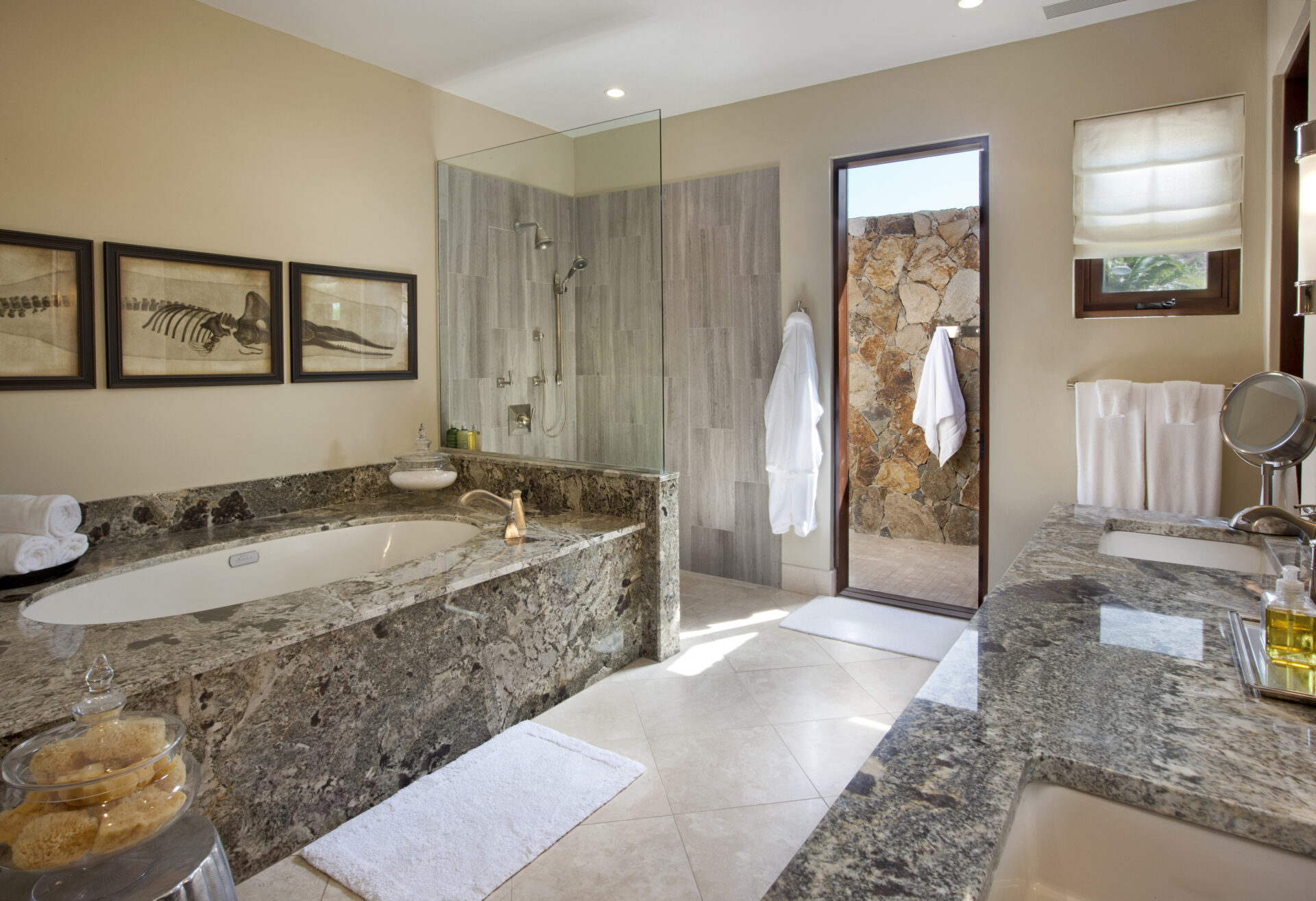 A luxurious bathroom at Bella Beach Villa featuring a large grey marble bathtub, a glass-enclosed shower, and a stone wall accent, along with double sinks with granite countertops. Three framed fish skeleton prints hang on the wall, and plush white towels are neatly arranged.