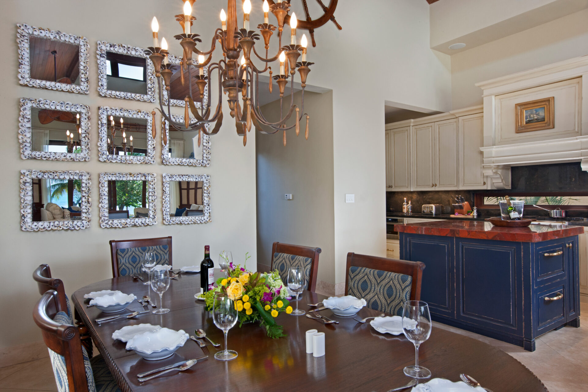 A dining room at Bella Beach Villa features a dark wooden table set for six, adorned with a floral centerpiece and wine bottle. A chandelier hangs above, illuminating the adjacent open kitchen with dark blue and white cabinets and a brown countertop. Nine wall mirrors in three rows decorate one wall.