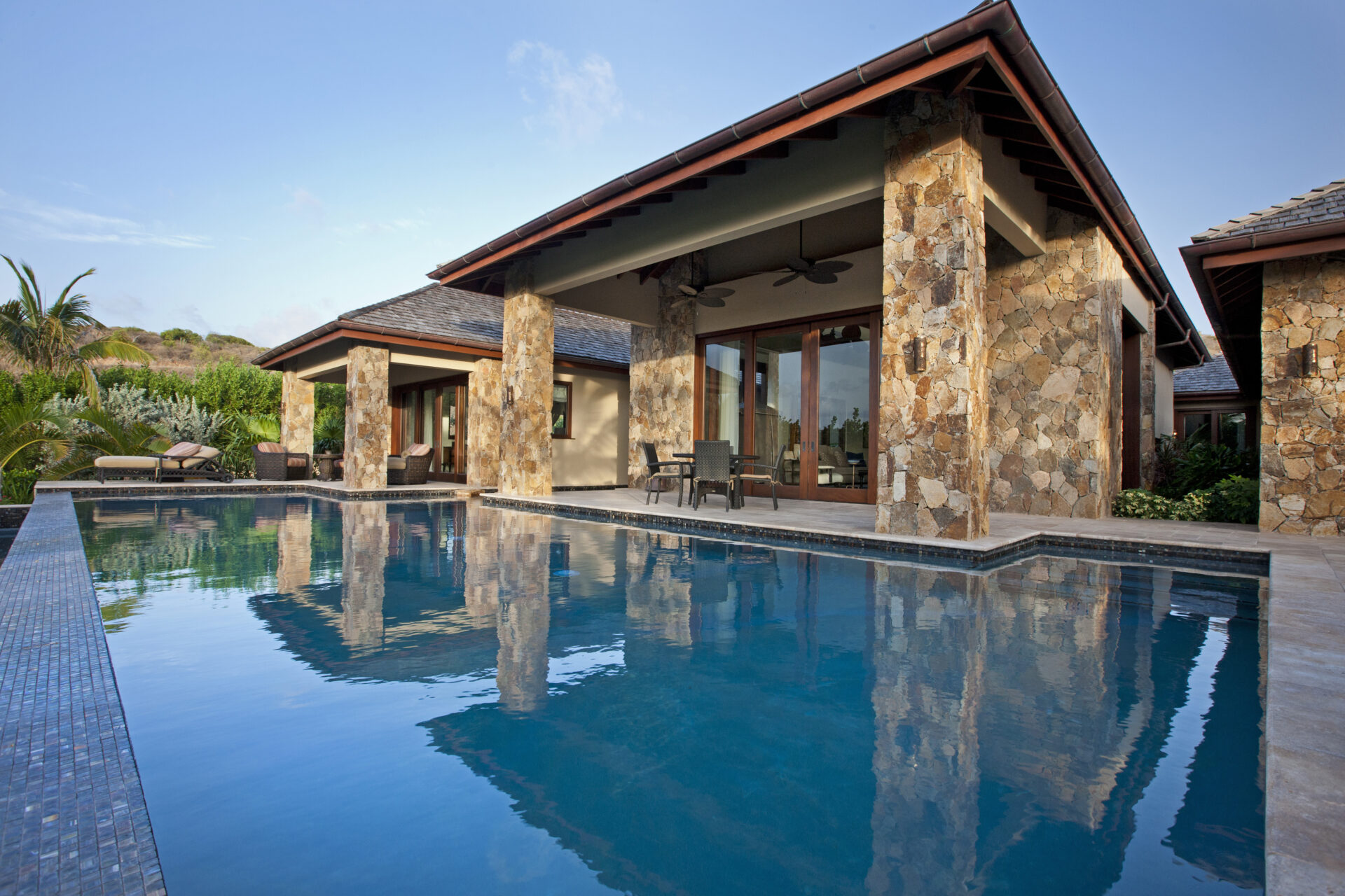 Bella Beach Villa is a luxurious house with stone pillars and large glass doors, situated behind an infinity pool. The contemporary design features a wooden roof and a spacious patio with outdoor furniture. The sky is clear, with lush greenery surrounding the property.