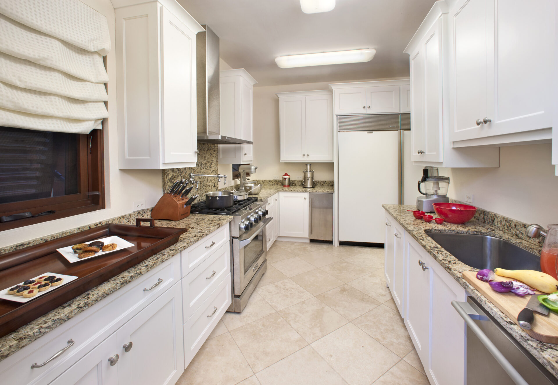 A modern kitchen at Bella Beach Villa features white cabinets, granite countertops, stainless steel appliances, and tiled flooring. A tray with food rests on the counter near the window with roman shades, while various fruits and utensils adorn the opposite counter.