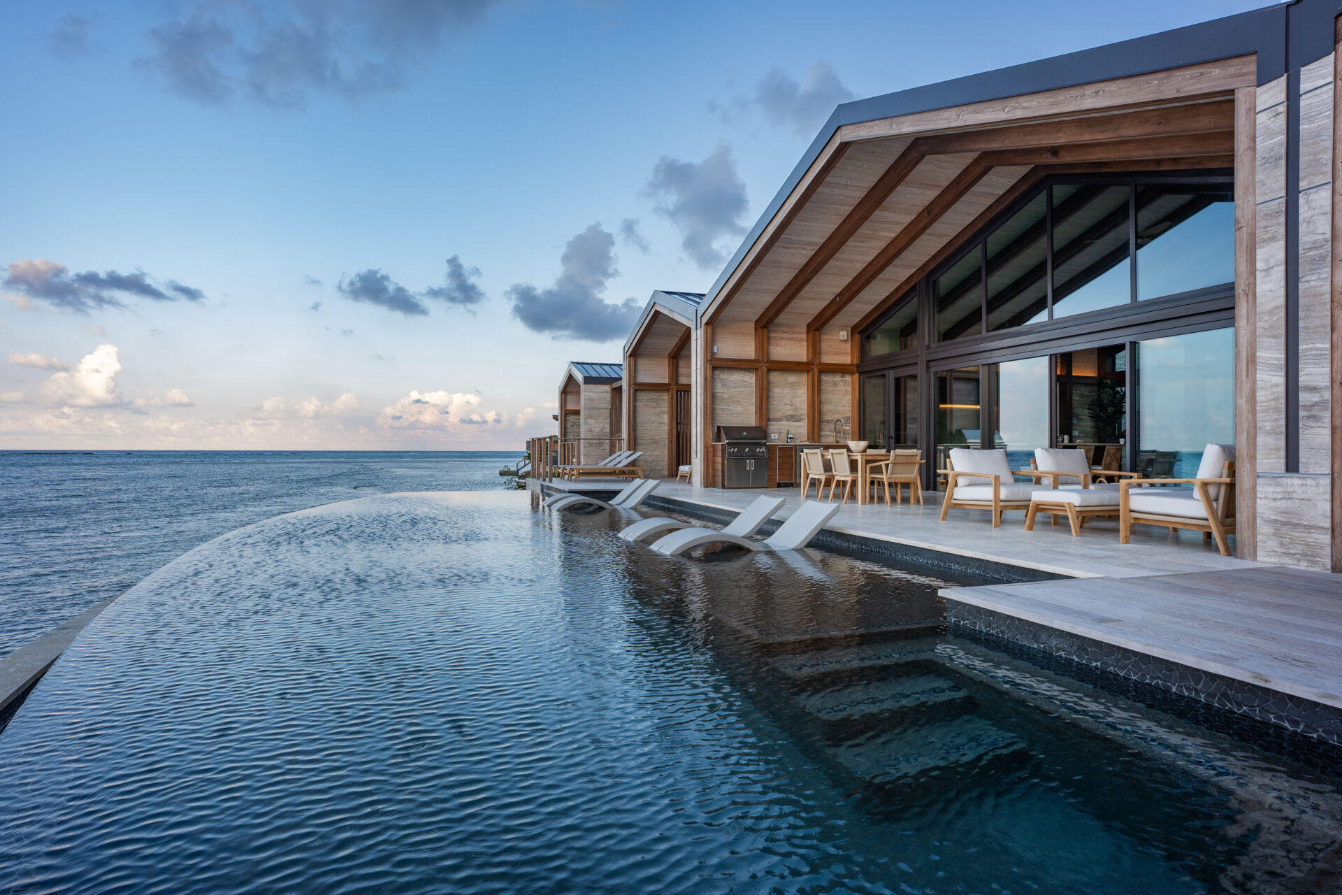 Ocean Villa: Modern oceanfront villa with large floor-to-ceiling windows, wooden beams, and an infinity pool stretching towards the sea. The patio area includes lounge chairs and a dining set, overlooking a serene ocean view under a partly cloudy sky.