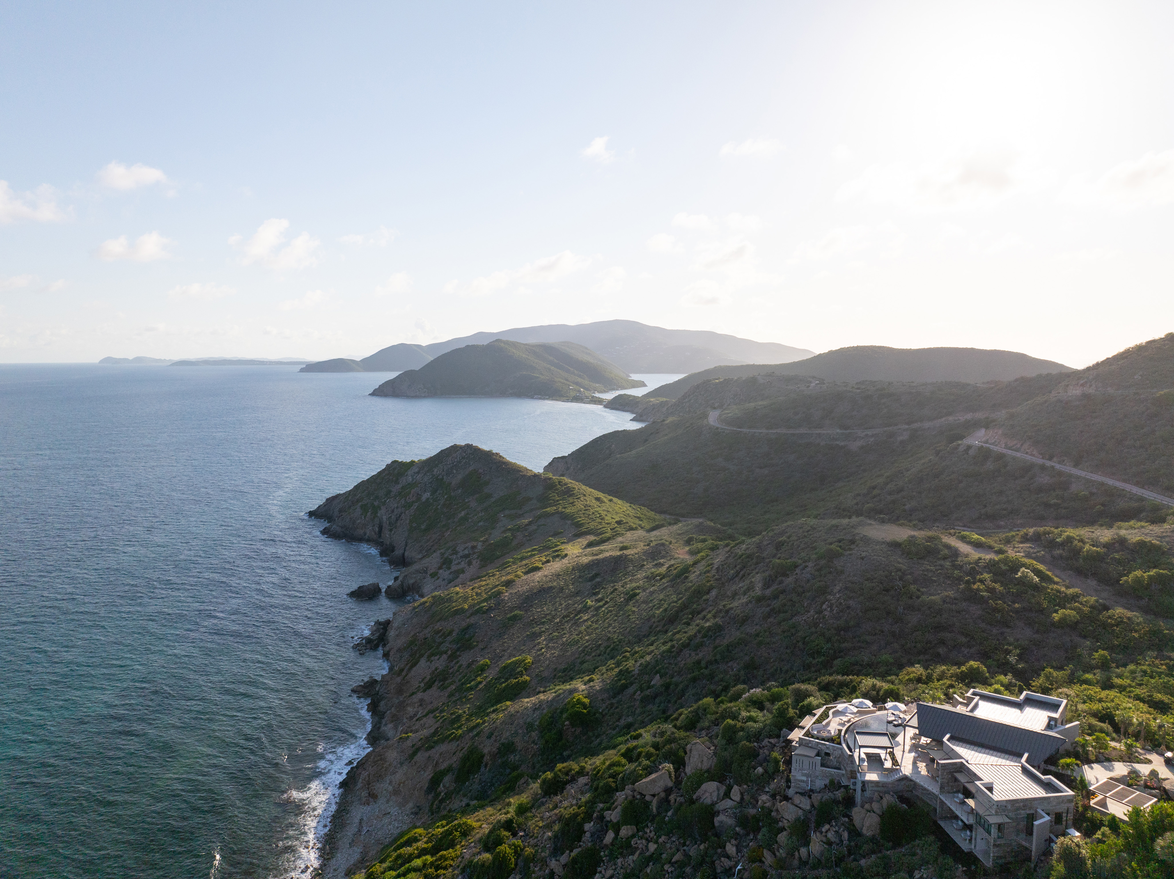 Aerial view of a rugged coastal landscape with lush green hills and Quintessa Villa, a modern, white luxury villa perched on a cliff overlooking the ocean. The coastline curves gently with rocky outcrops, and the sky is partly cloudy with the sun low on the horizon.