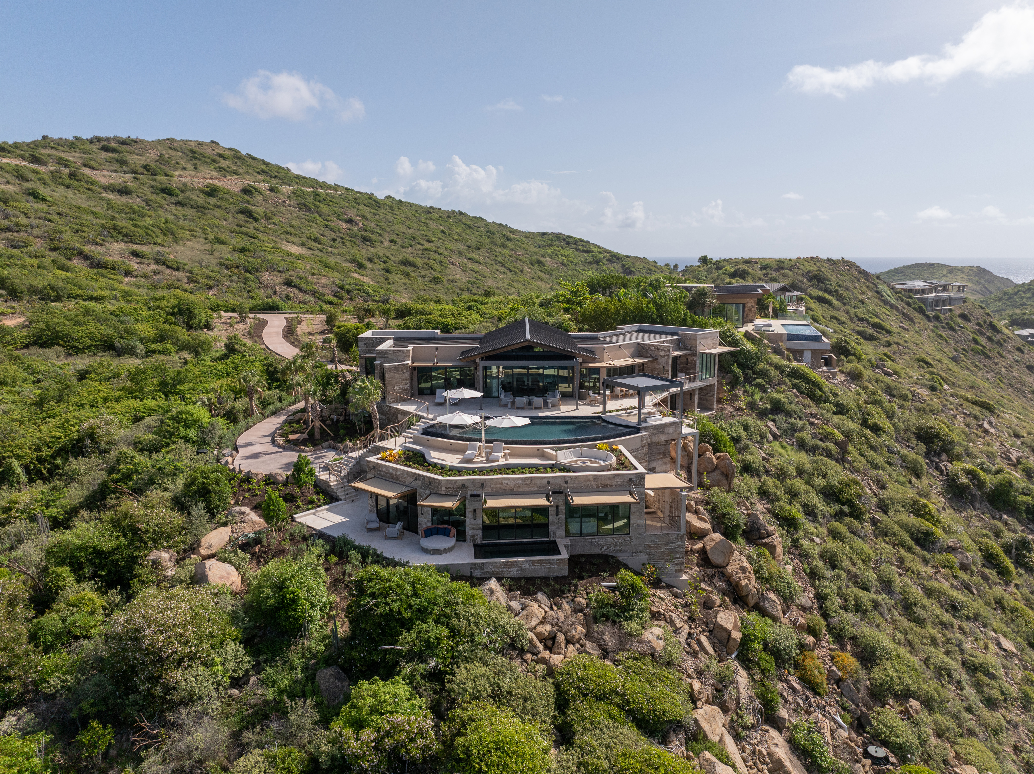 A modern, multi-level luxury home, Quintessa Villa boasts large glass windows and balconies nestled on a lush, green hillside overlooking the ocean. The house features an expansive outdoor pool area with umbrellas and sun loungers, surrounded by rolling hills and scenic views.