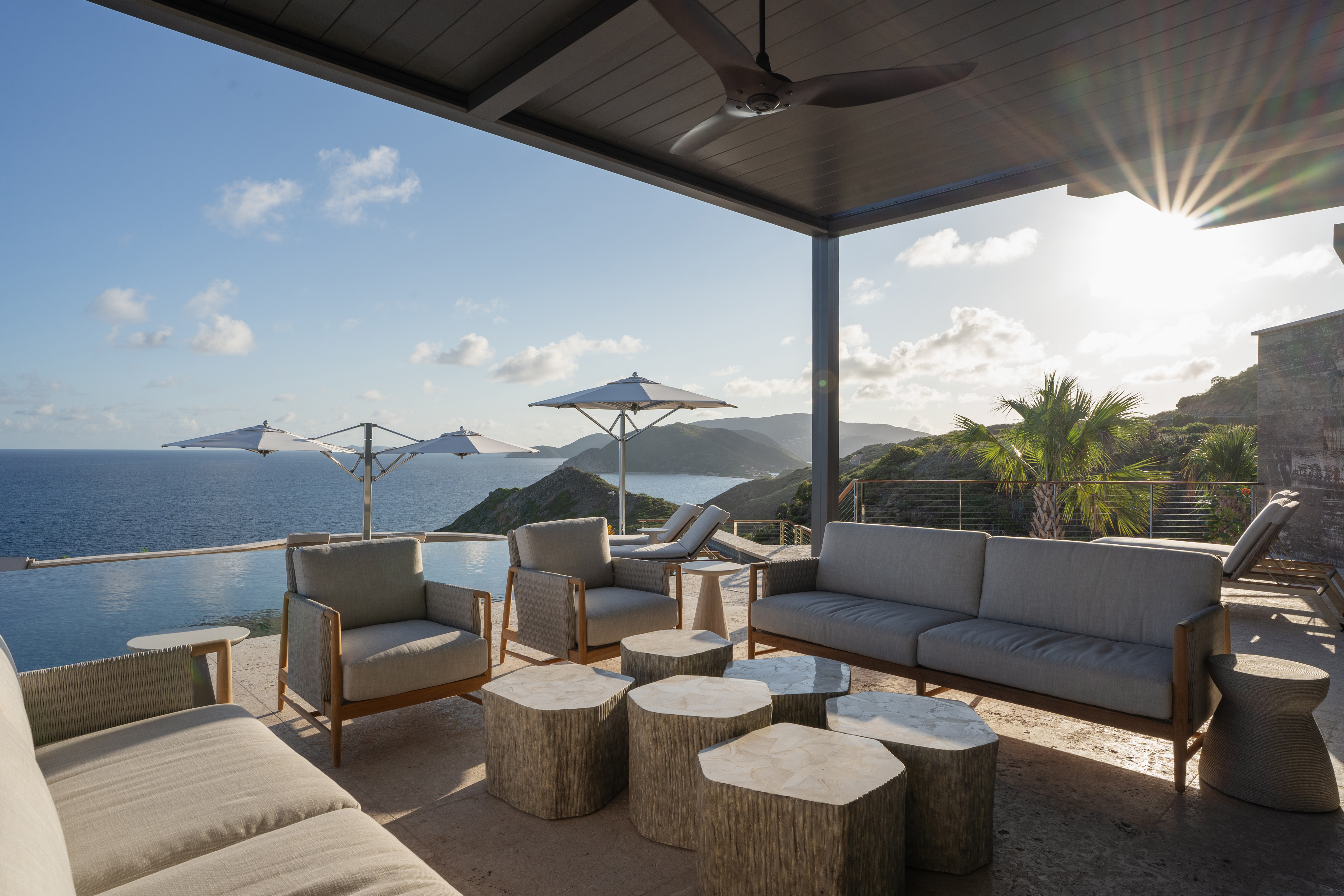 A spacious outdoor patio at Quintessa Villa features modern wooden furniture, including cushioned sofas and circular tables, overlooking a scenic ocean view. Large umbrellas provide shade, and the setting sun casts a warm glow over the peaceful coastal landscape.