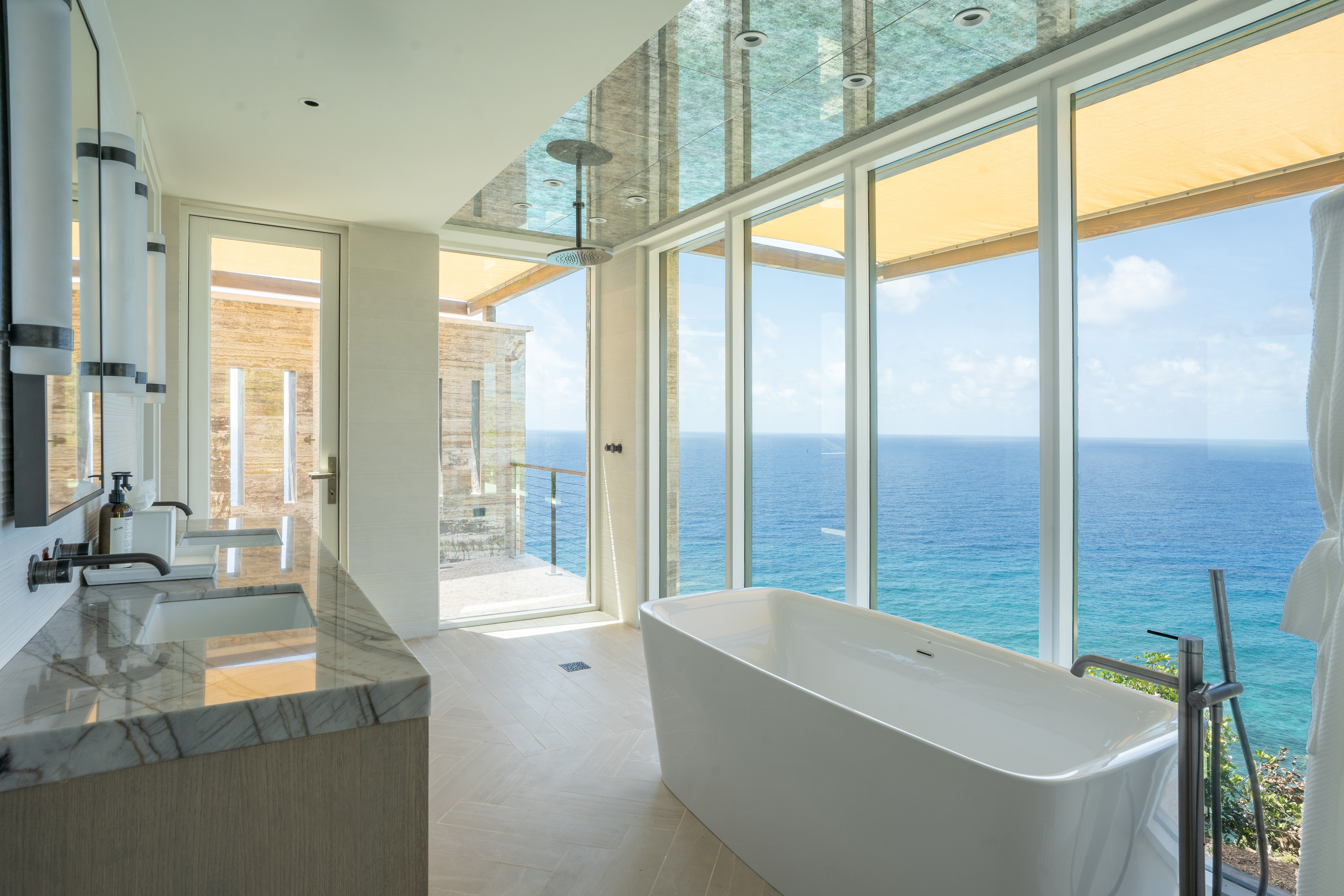 A modern bathroom at Quintessa Villa boasts a marble-topped double sink vanity on the left, a freestanding bathtub in the center, and floor-to-ceiling windows showcasing a stunning ocean view. The ceiling features skylights, and the room is filled with natural light.