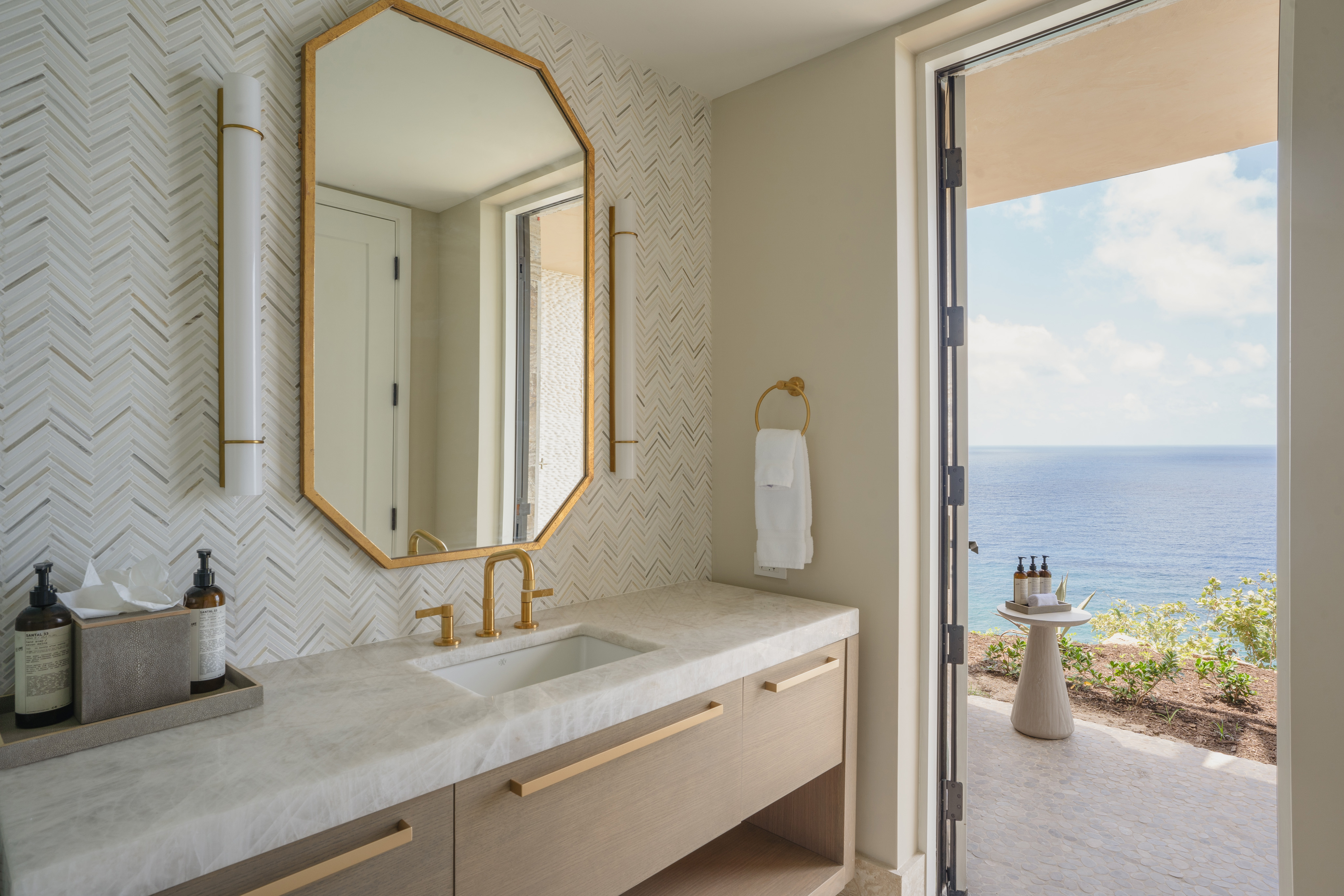 Modern bathroom with beige and white herringbone-patterned walls, a beige vanity with a marble countertop, a gold-framed mirror, and gold faucet. An open door reveals a patio with a small table overlooking an ocean view at the luxurious Quintessa Villa vacation rental.