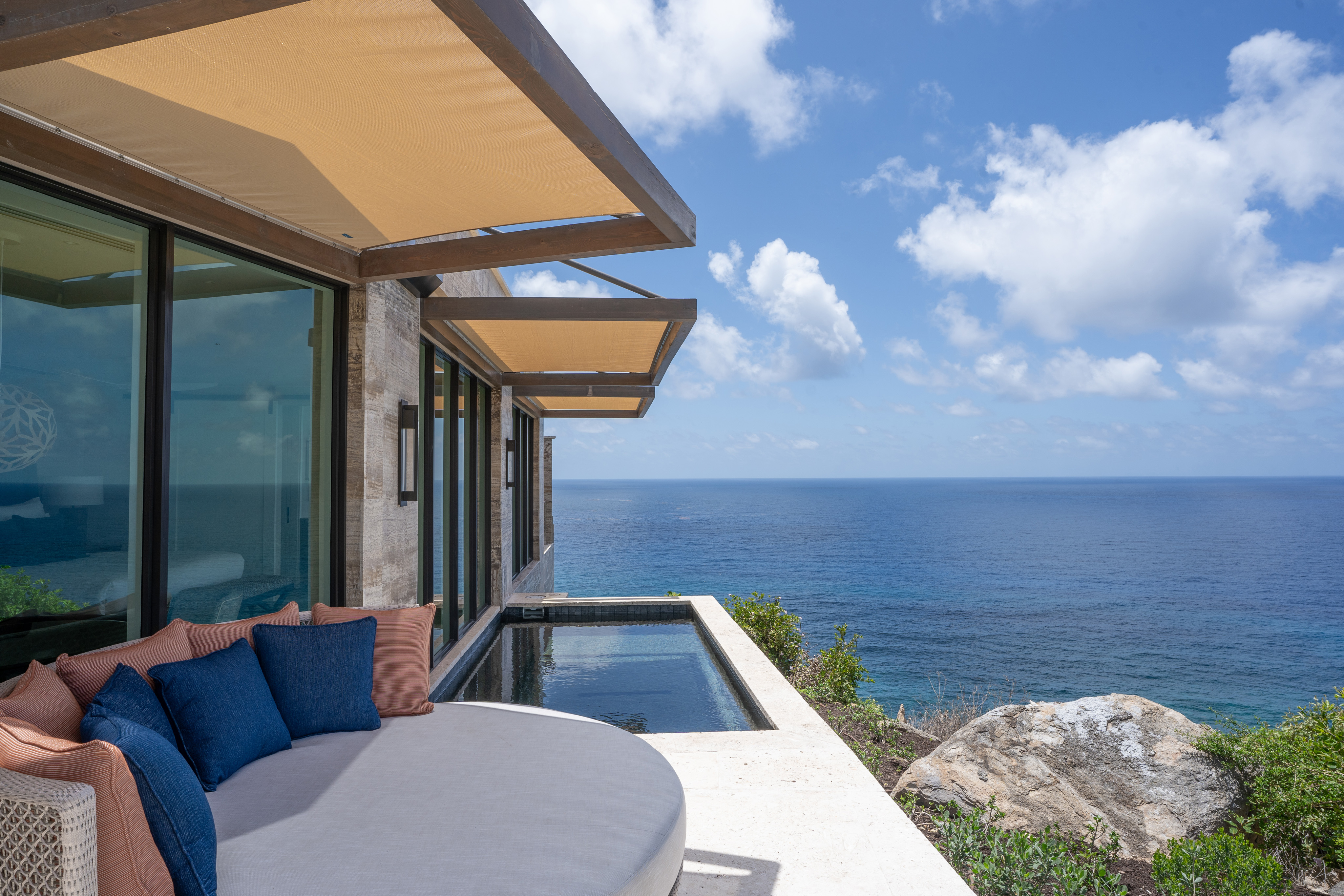 Quintessa Villa: A modern seaside haven featuring beige awnings, glass windows, and a round cushioned lounge area adorned with blue and beige pillows. The patio boasts a small infinity pool overlooking the vast blue ocean, all set against rocky cliffs and lush greenery in a stylish and serene setting.