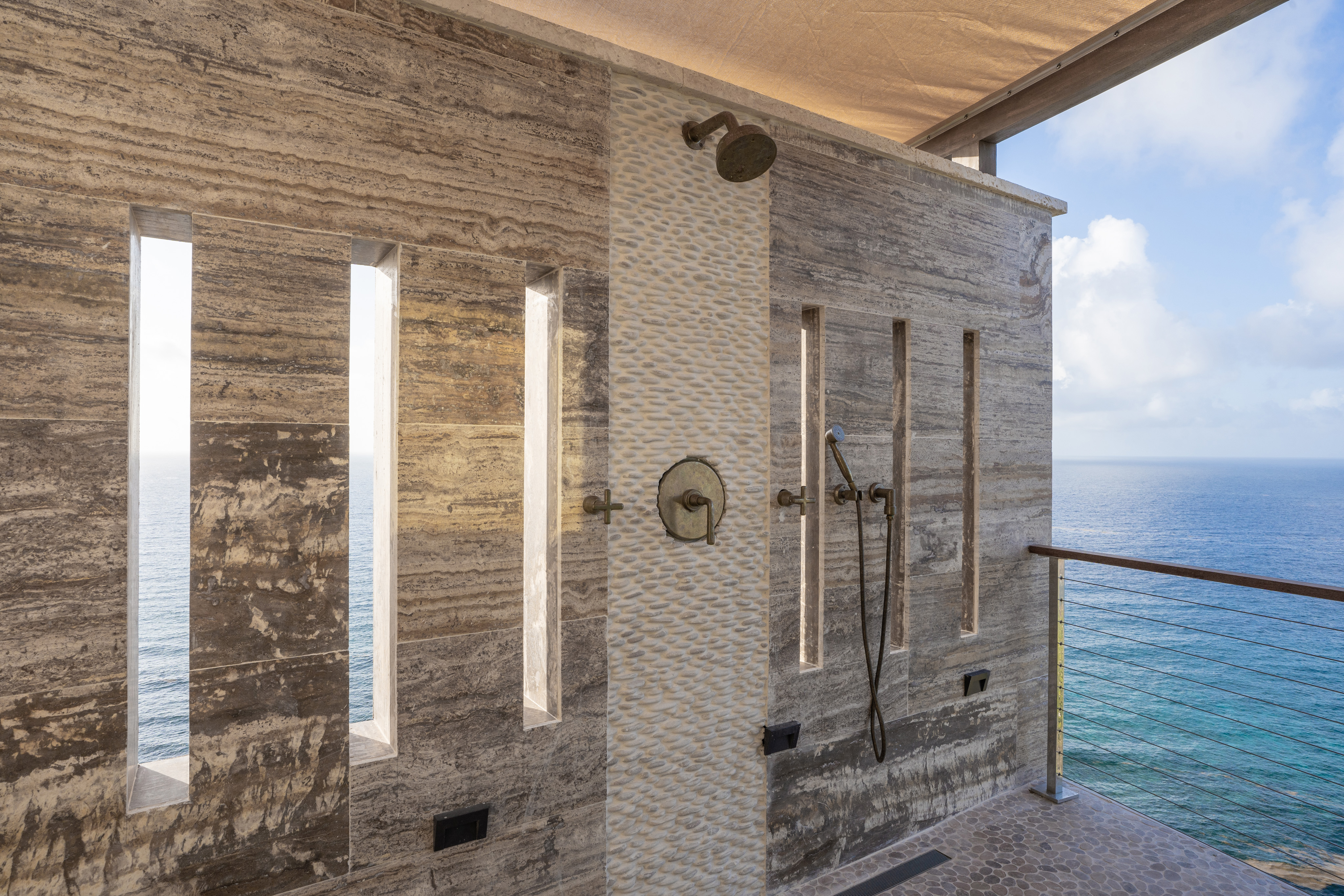 An outdoor shower at Quintessa Villa, with vertical slits in a stone wall, offers a stunning view of the ocean. The shower has both overhead and handheld showerheads and is positioned on a deck with a clear glass railing, allowing for an unobstructed sea view.