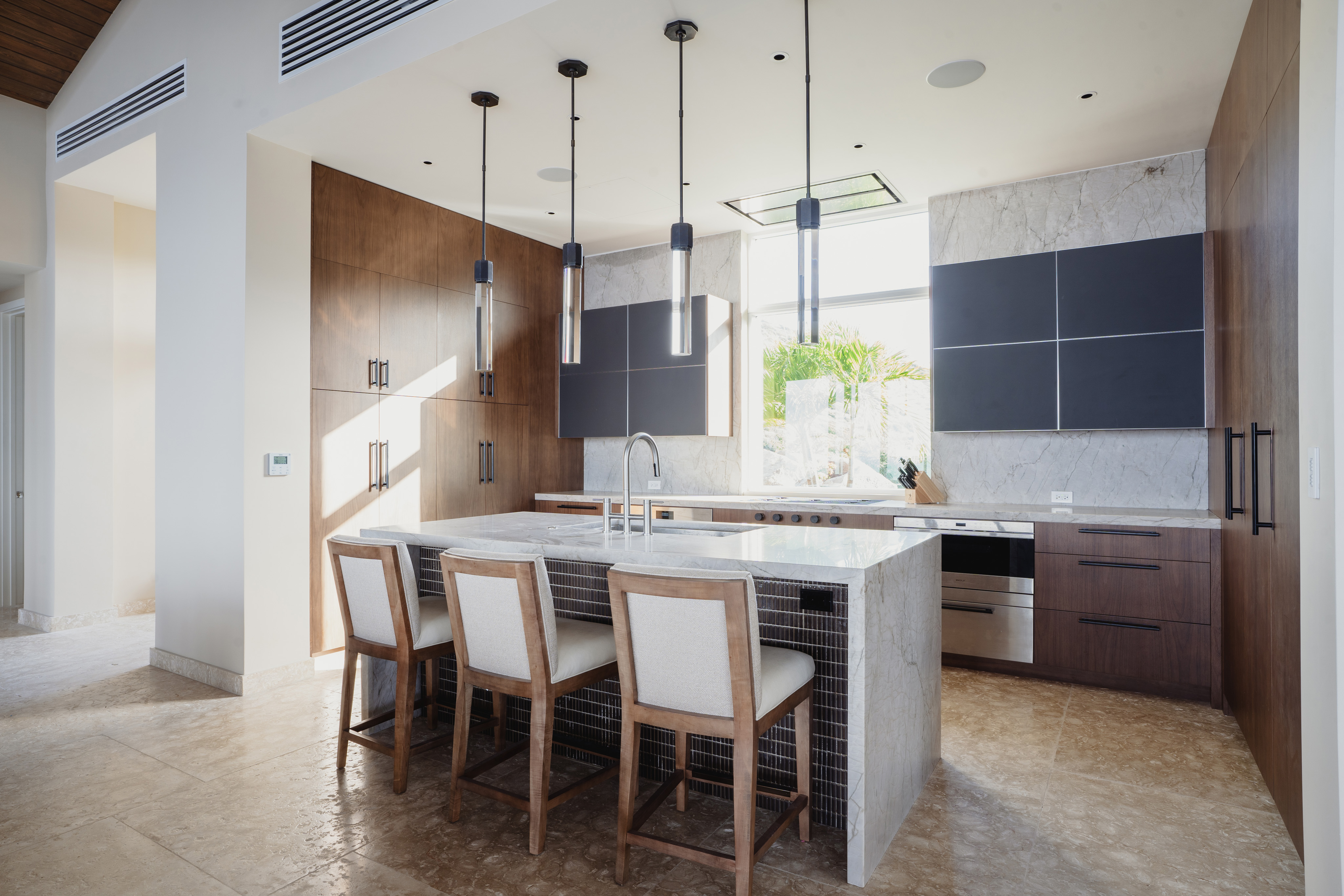 The Quintessa Villa features a modern kitchen with marble countertops and island, light wood cabinets, black upper cabinets, and stainless steel appliances. Three gray pendant lights hang above the island, which has three wooden chairs with cushioned seats. Natural light illuminates the space.