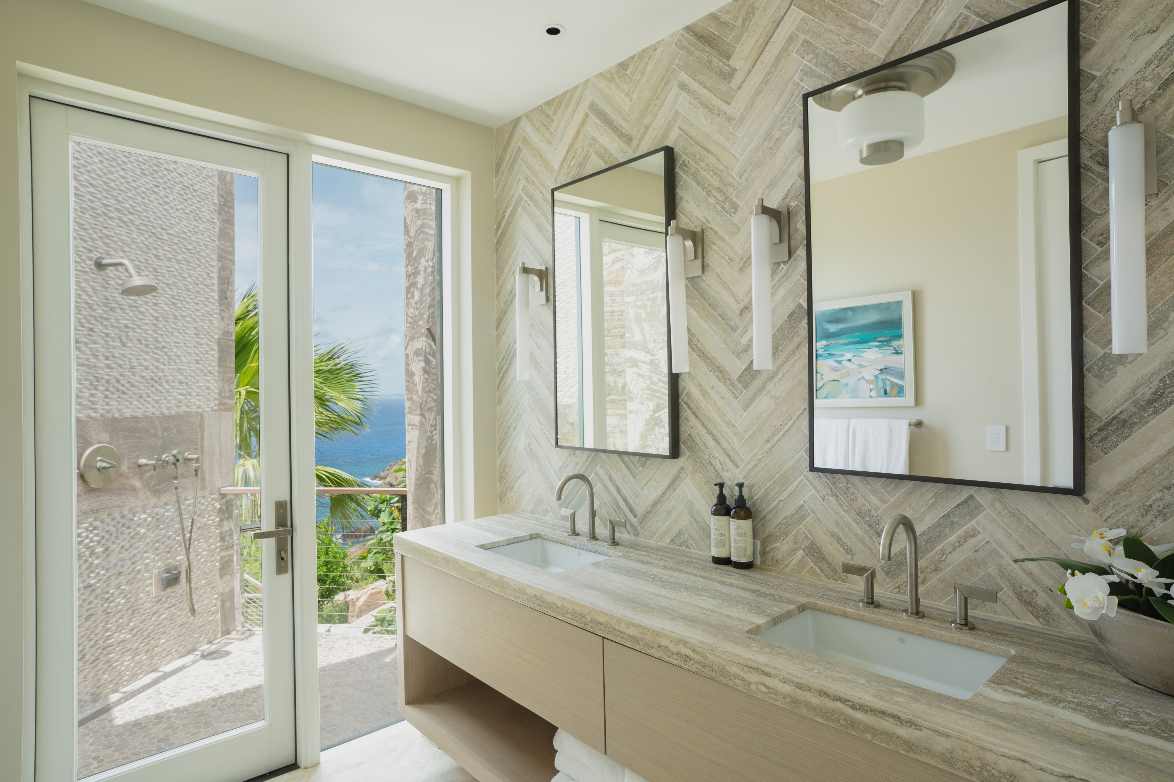 A modern bathroom in the luxurious Quintessa Villa features a large glass door opening to a scenic outdoor view with a glimpse of the ocean and tropical foliage. The interior includes a dual-sink vanity with a marble countertop, twin mirrors, sleek lighting fixtures, and decorative accents. Enjoy the ultimate vacation rental experience here.