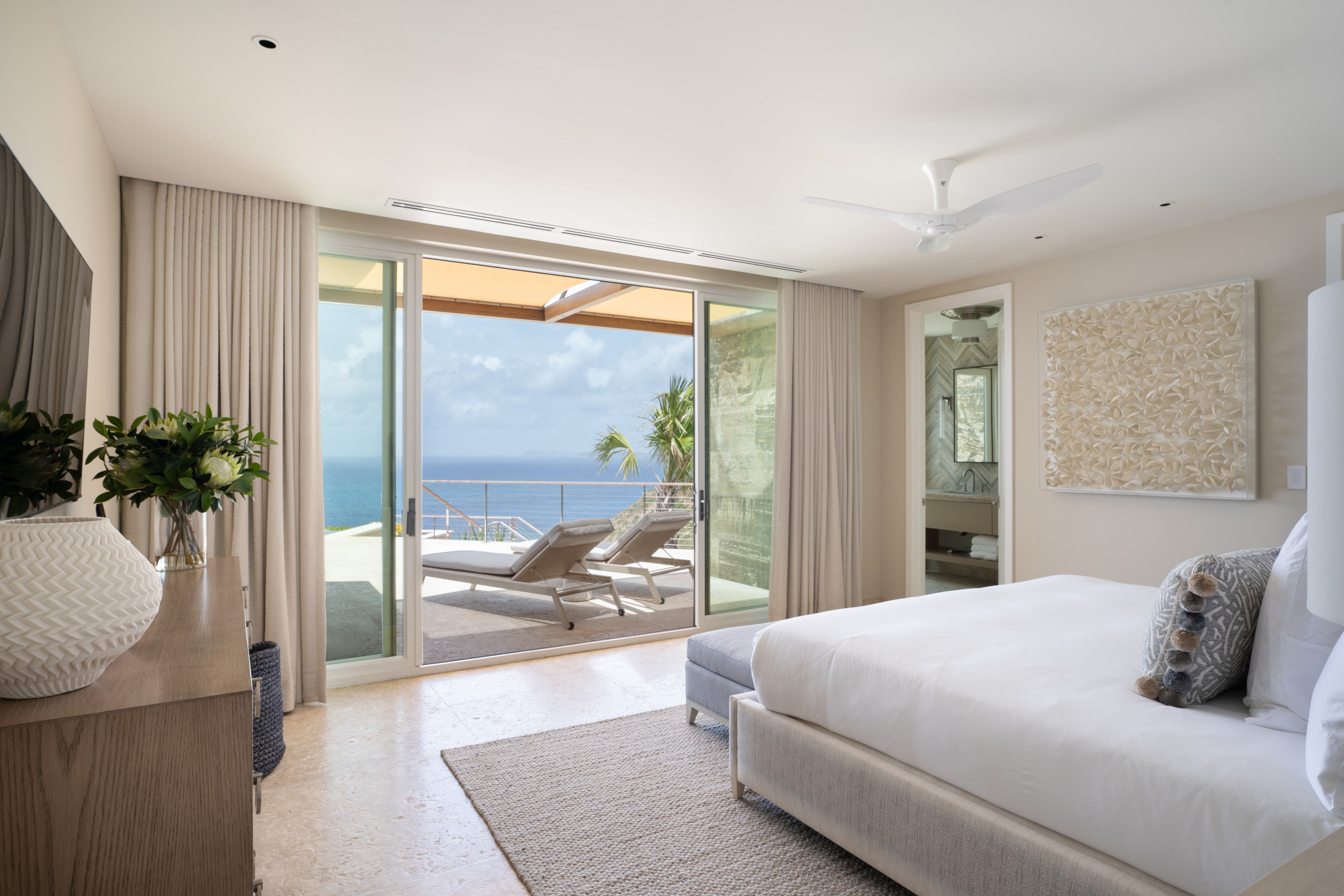 A bright, modern bedroom at Quintessa Villa with light-colored furnishings and decor defines elegance. The room features a large bed, a dresser with a vase of flowers, and floor-to-ceiling sliding glass doors that open to a patio with lounge chairs overlooking a stunning ocean view.