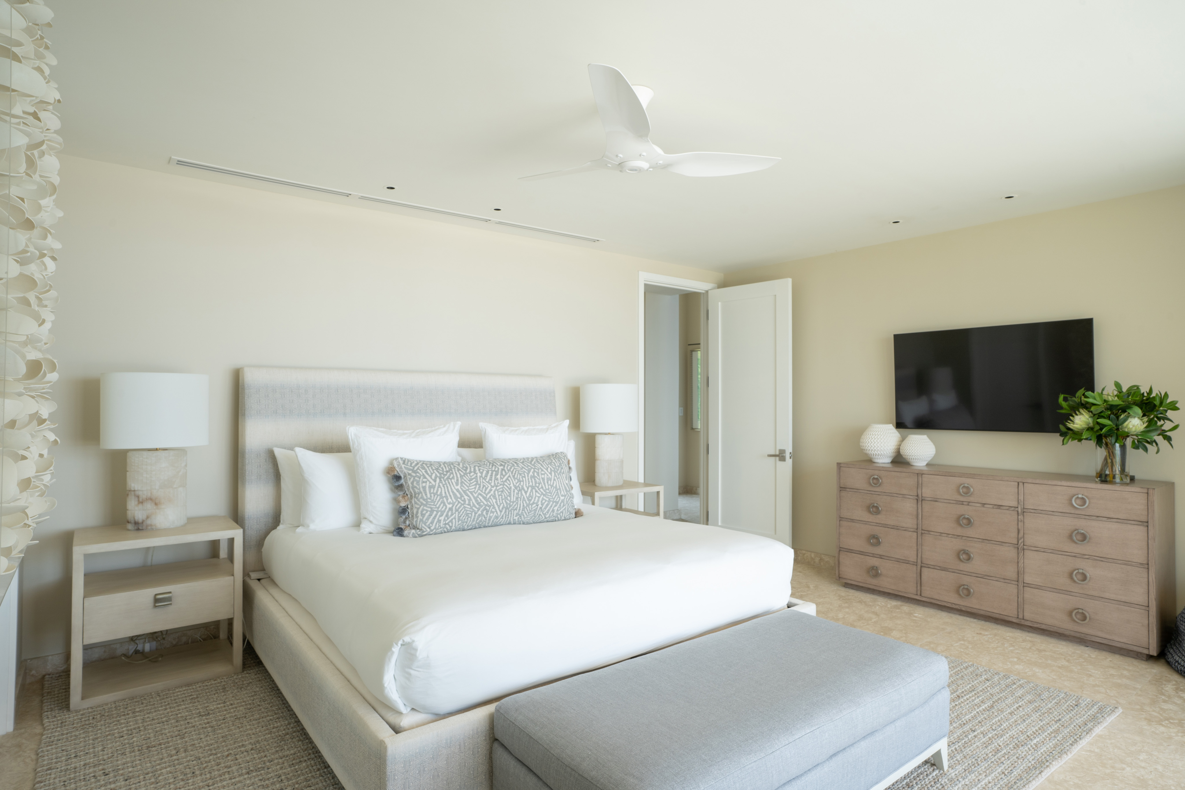 A modern, beige-themed bedroom at Quintessa Villa features a large bed with white linens and multiple pillows, flanked by two nightstands with matching lamps. The room includes a ceiling fan, a flat-screen TV mounted on the wall, a dresser with decor, and a cushioned bench at the foot of the bed.