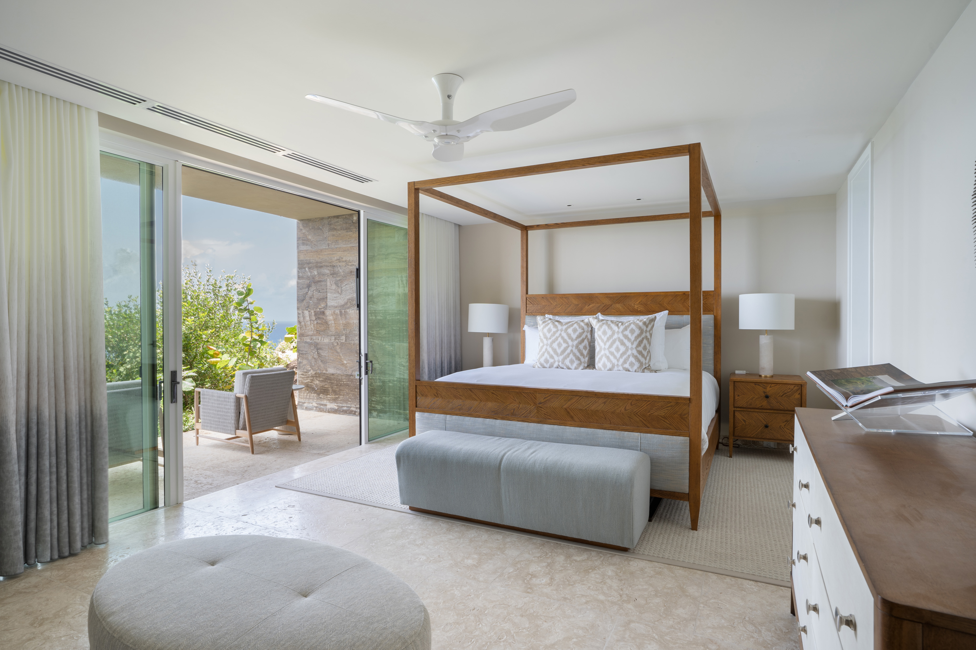 A modern bedroom in Villa Quintessa features a wooden four-poster bed, white bedding, and decorative pillows. A light blue bench sits at the foot. There is a dresser on the right and a seating area outside on the balcony, accessible through sliding glass doors. The room is bright and airy.