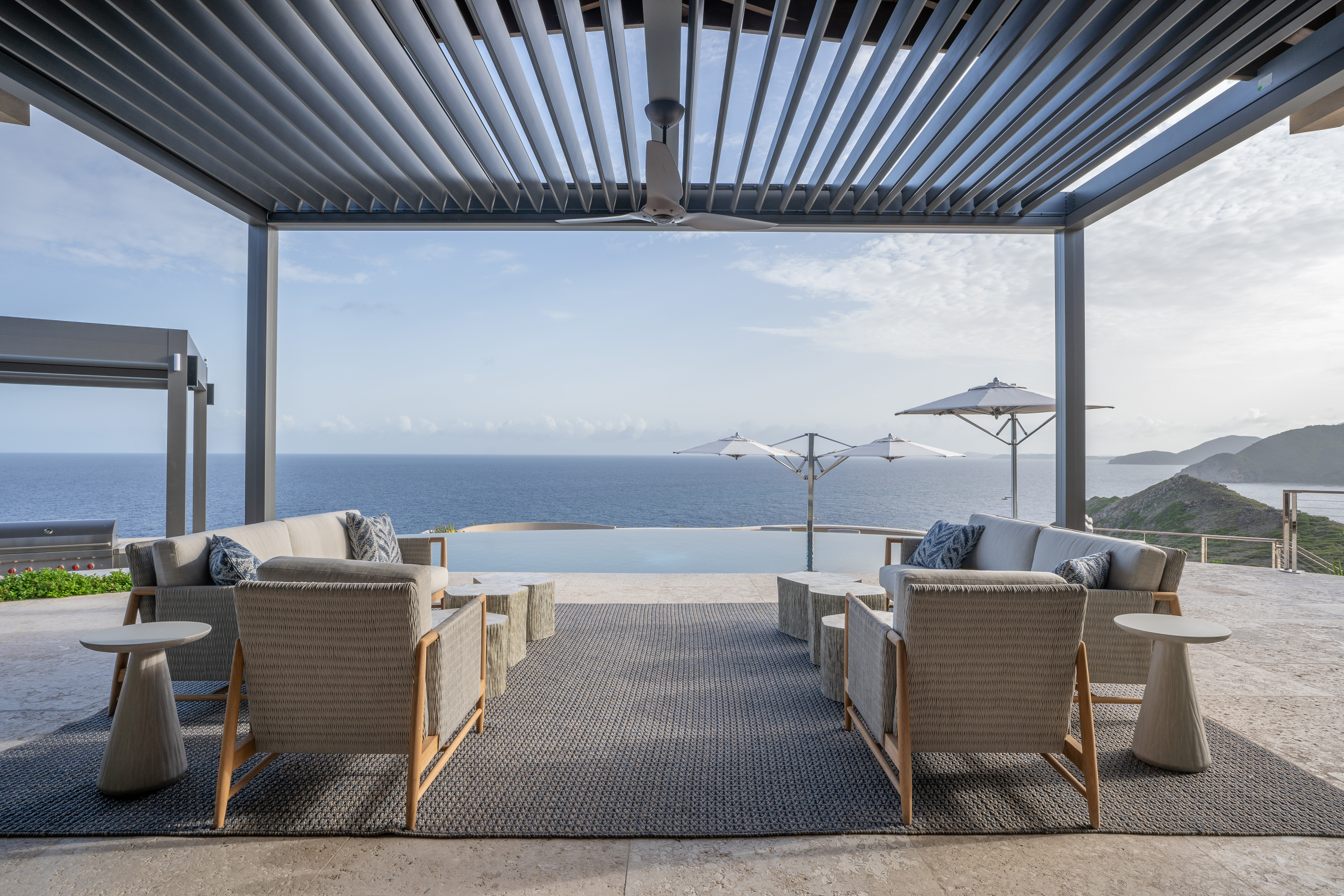 Outdoor terrace at Quintessa Villa overlooking the ocean with comfortable seating arrangements, including armchairs and sofas around a coffee table. The area is shaded by a pergola, and there are umbrellas in the distance. The serene scene includes clear blue skies and rolling hills.