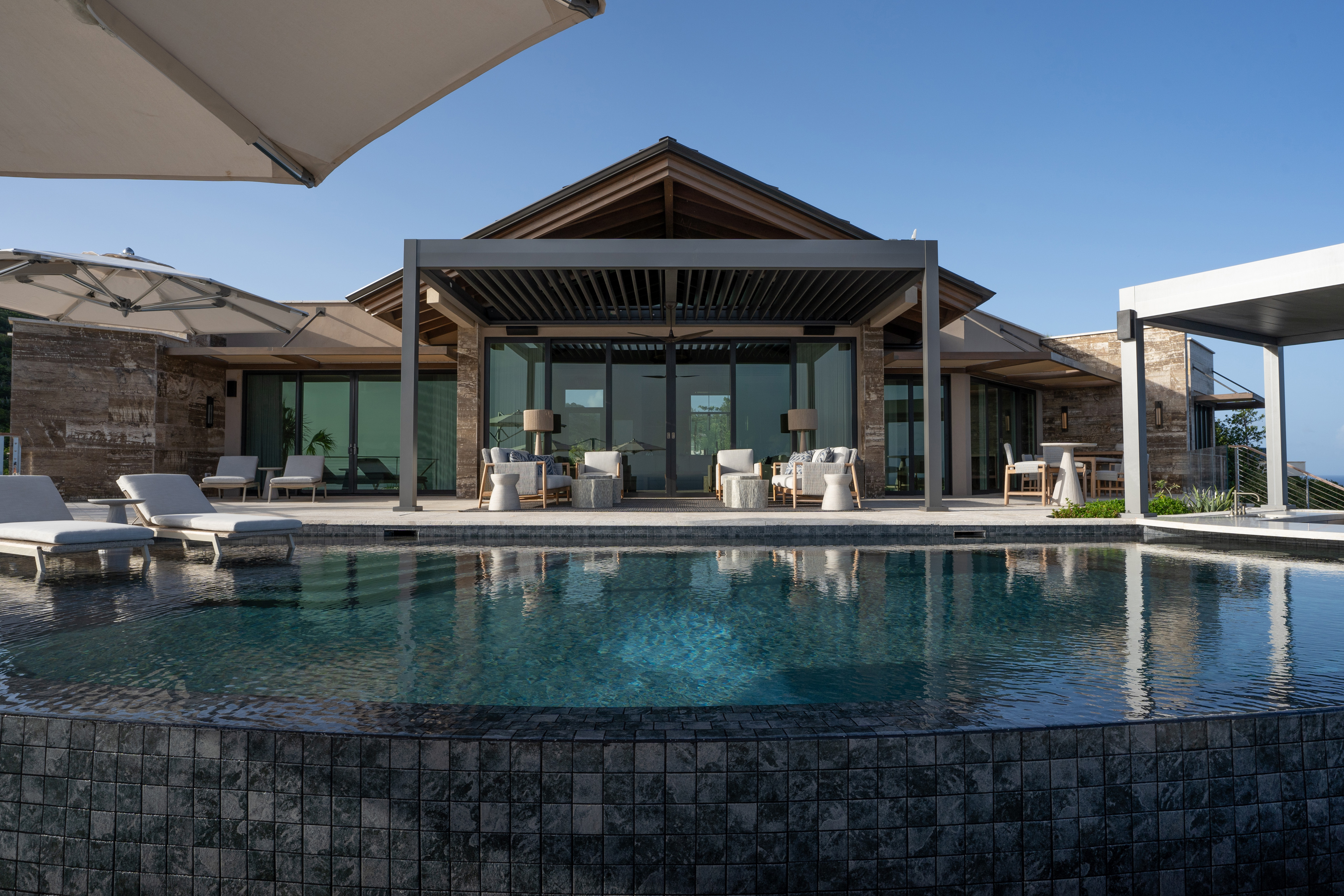 The Quintessa Villa is a luxurious modern house with a large terrace featuring plush outdoor seating and a stunning infinity pool that overlooks the scenic landscape. The house has extensive glass windows and a contemporary architectural design, surrounded by clear blue skies.