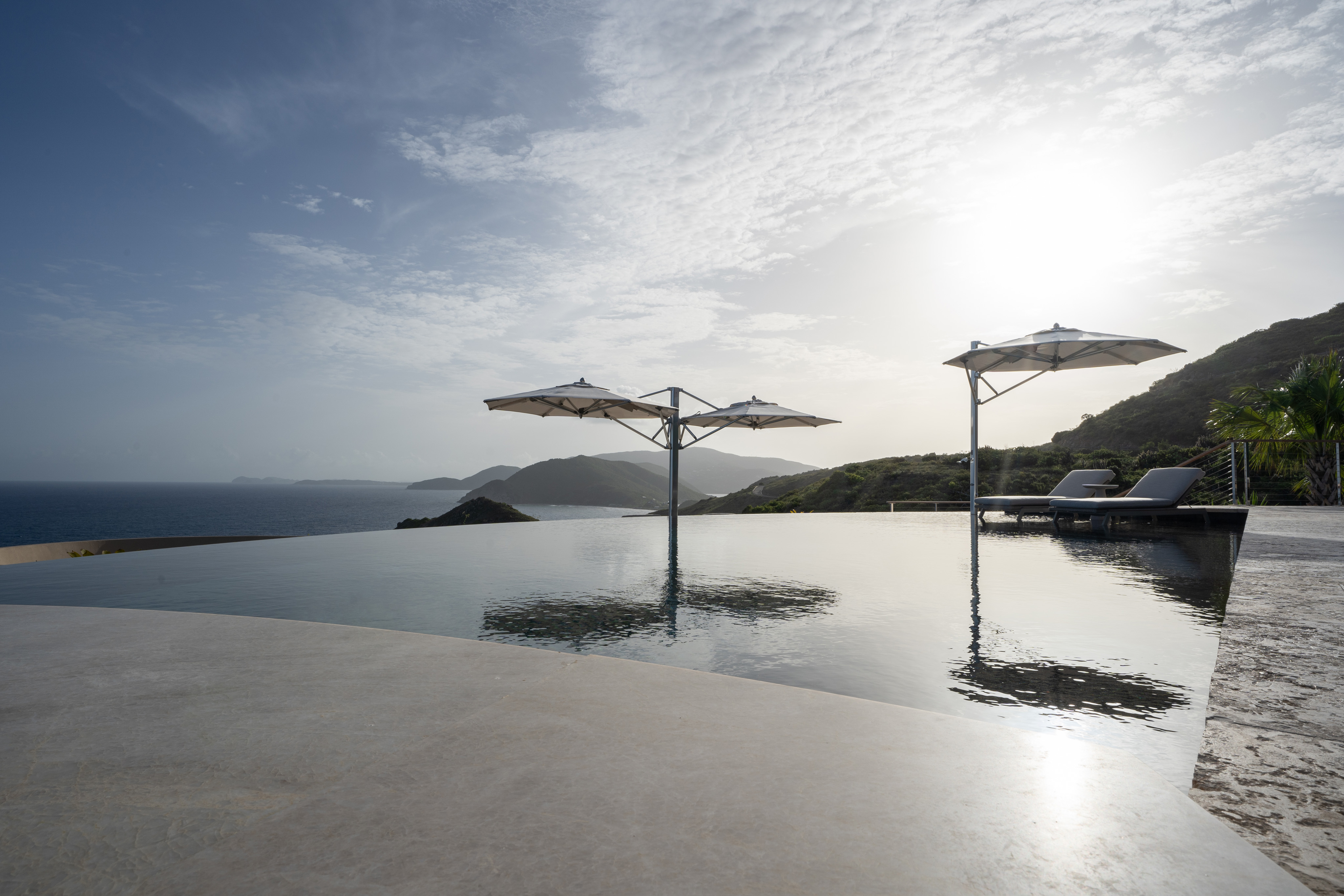 At Quintessa Villa, an infinity pool reflects the sky under two large white umbrellas, with lounge chairs positioned underneath. The pool overlooks a scenic coastal and mountainous landscape, with sunlight casting a glow over the horizon, partially obscured by clouds.