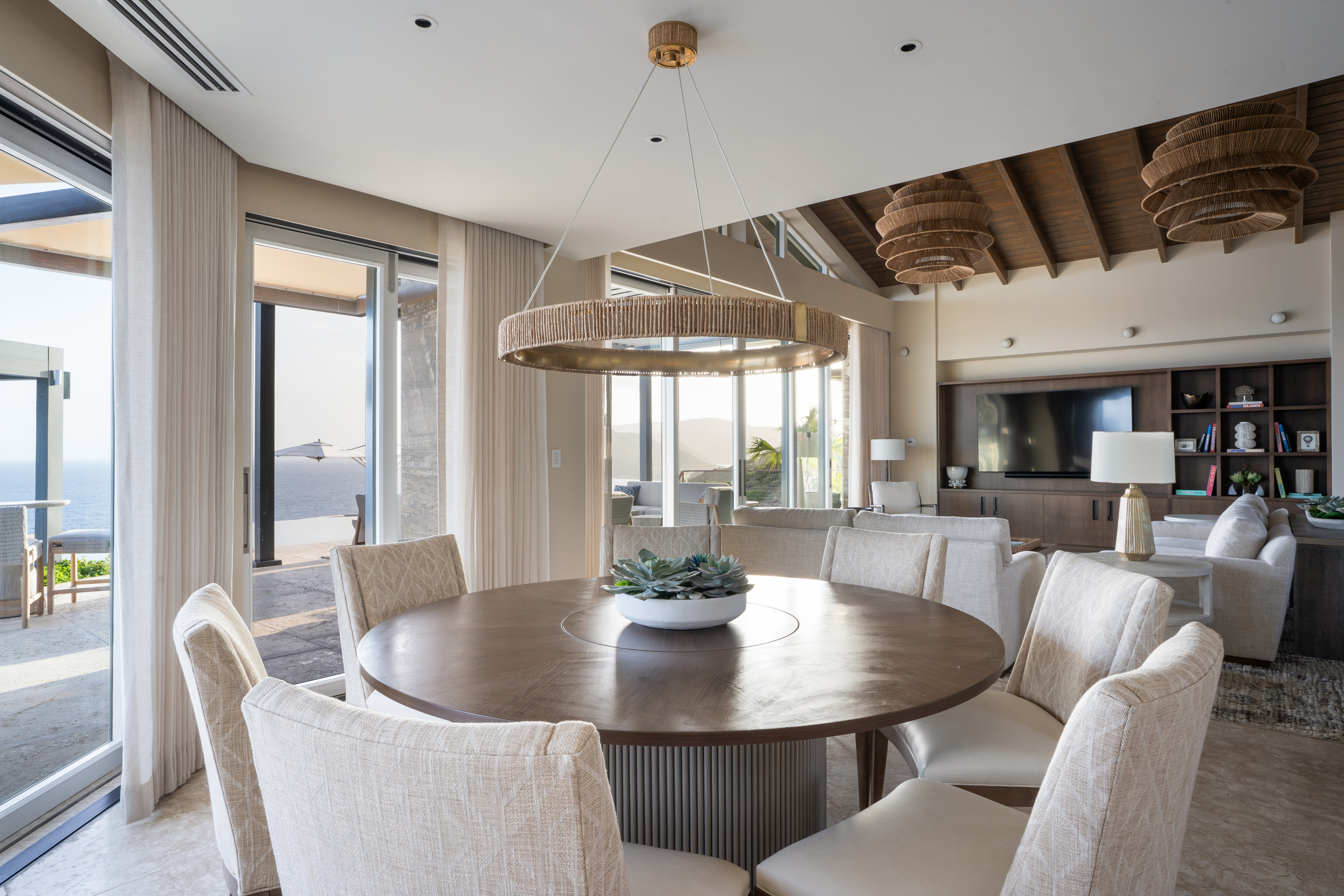 A modern dining room in the Quintessa Villa features a round wooden table surrounded by six upholstered chairs. A large, circular pendant light hangs above, while windows offer views of the ocean and patio. The background reveals a cozy living area with a TV and shelves, perfect for your vacation rental.