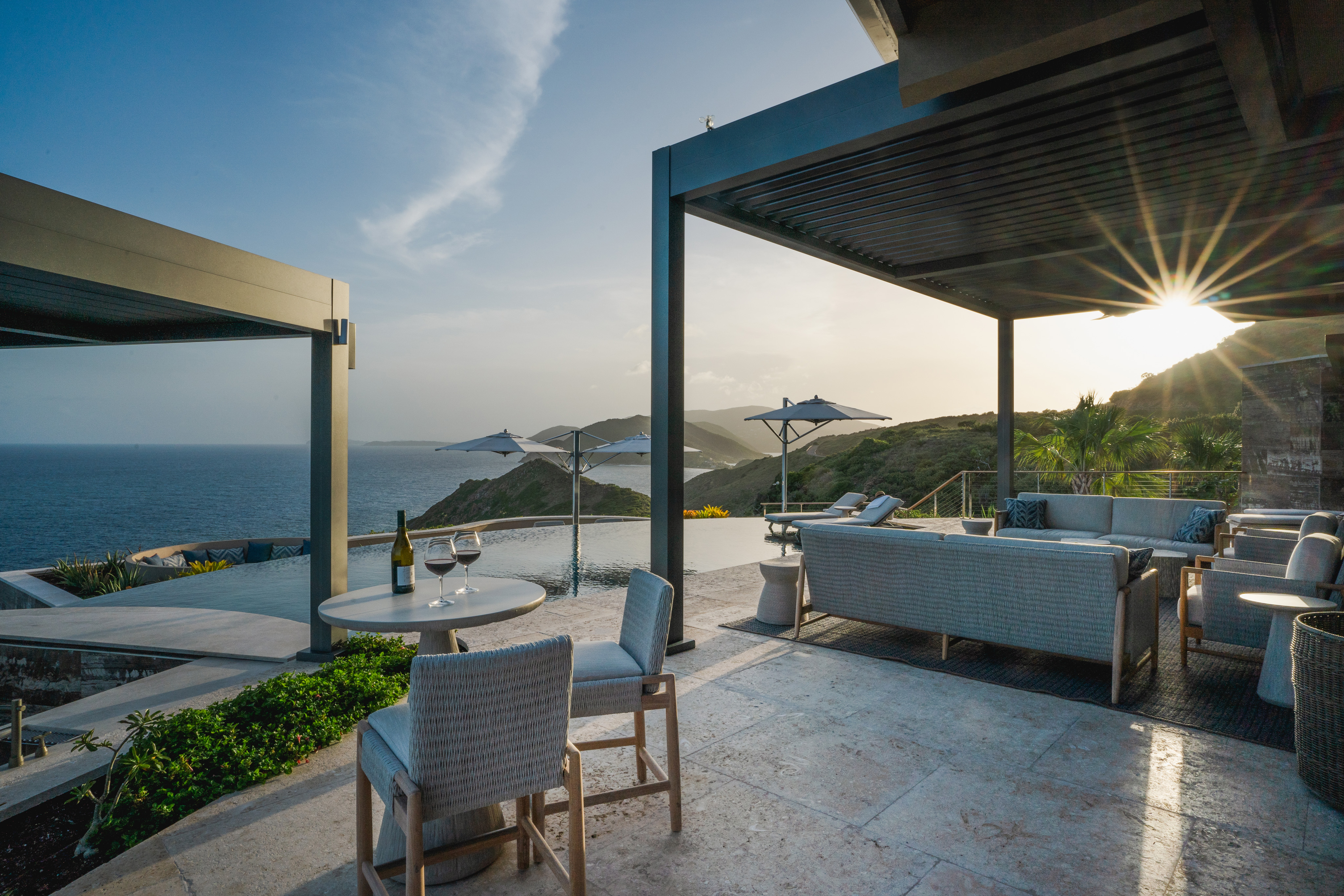 Outdoor patio with wicker furniture and a small table set for two, overlooking an infinity pool and ocean at the exquisite Quintessa Villa. Sun rays peek through a pergola's slatted roof; umbrellas and distant hills enhance the serene, luxurious atmosphere. A bottle of wine and two glasses sit on the table.