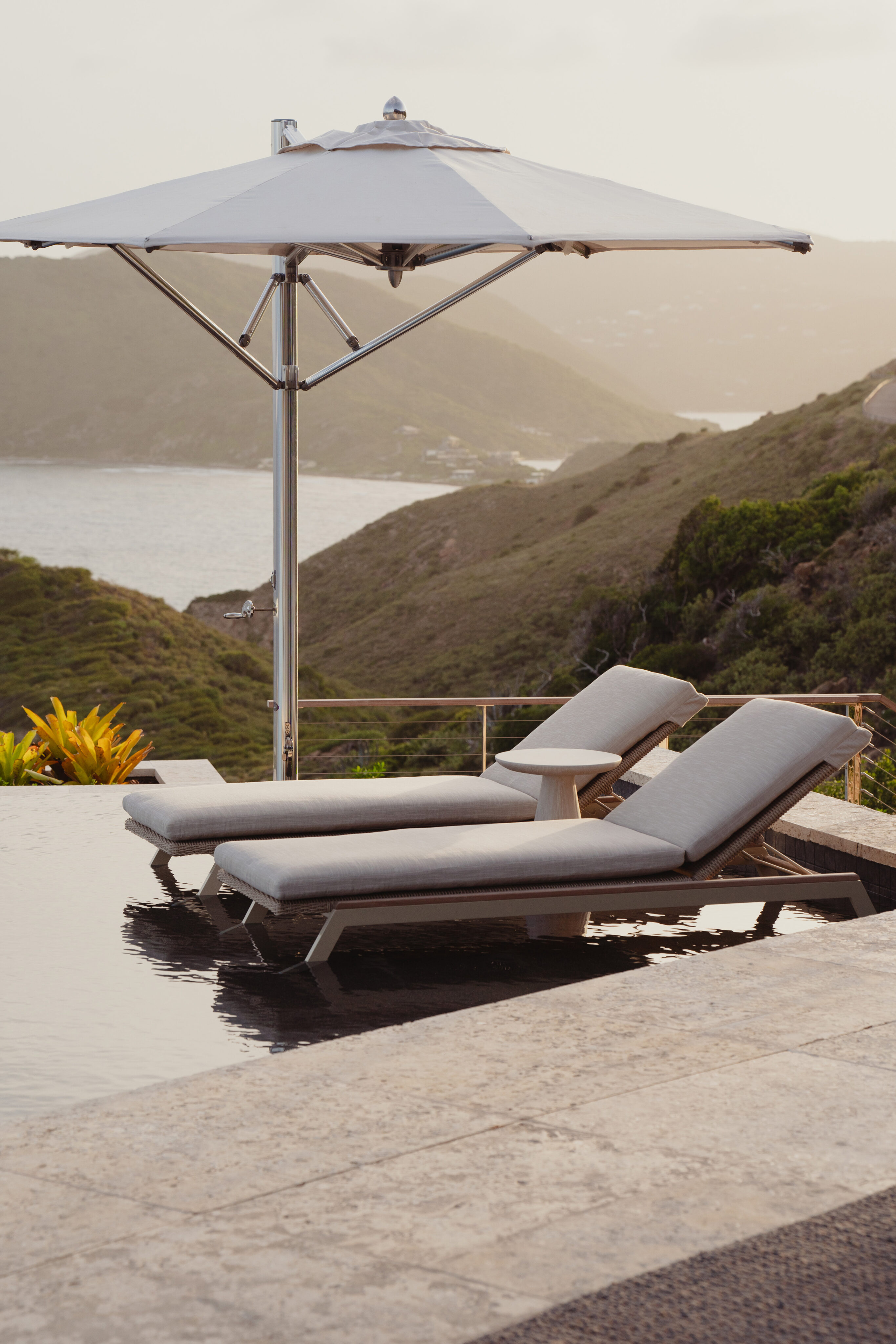 Two lounge chairs with gray cushions sit next to a reflective infinity pool at Villa Quintessa, shaded by a large white umbrella. The pool overlooks a scenic view of rolling green hills and a distant, tranquil body of water under a cloudy sky.