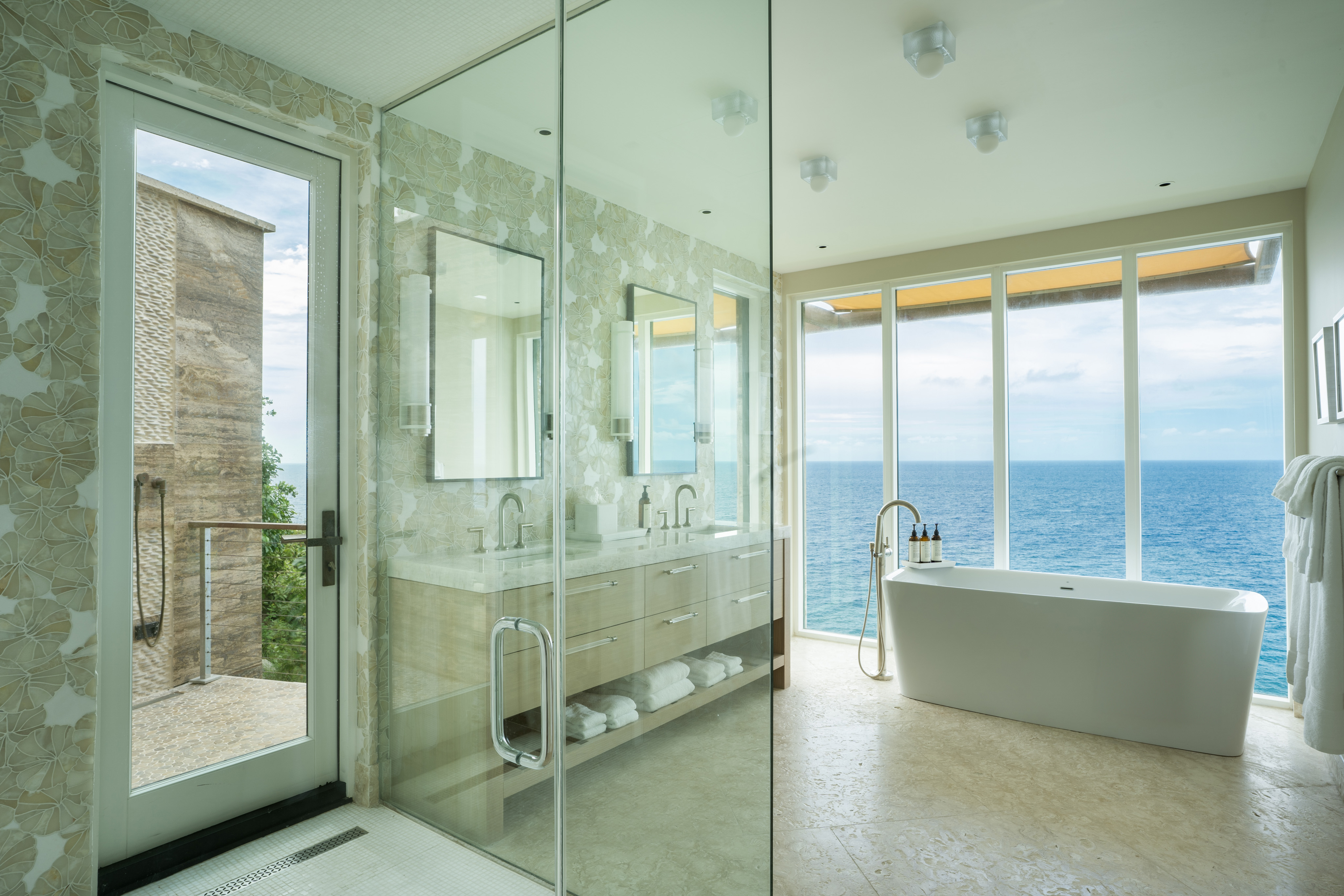 A modern bathroom in the luxury villa, Quintessa Villa, boasts a large window offering an ocean view. It features a glass-enclosed shower, a white freestanding tub, dual sinks with a large mirror above, and light-colored cabinets. Outside the window, part of the building and the sea are visible.