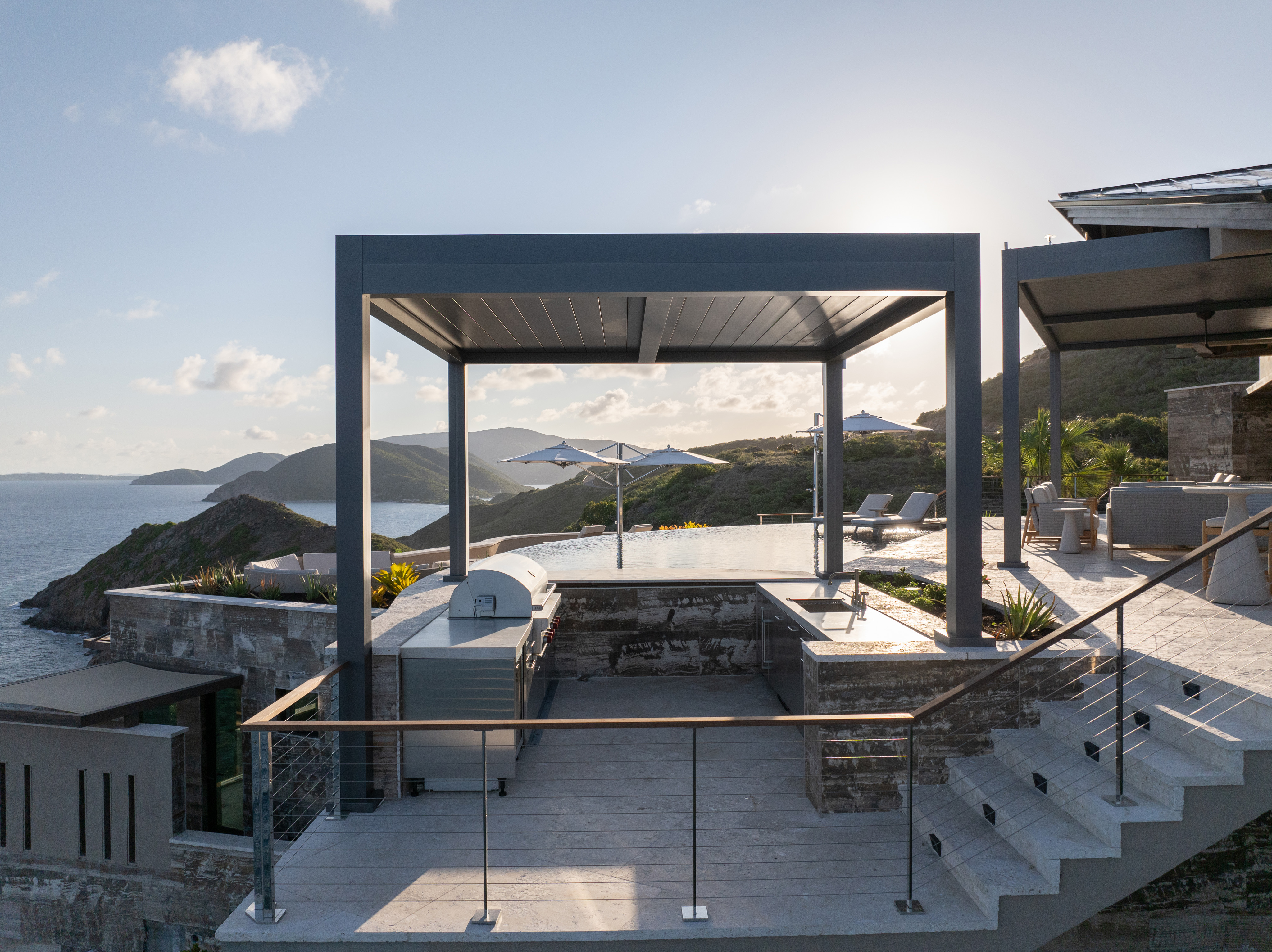 The Quintessa Villa boasts a modern rooftop patio with a pergola, outdoor kitchen, and an infinity pool overlooking a serene coastal landscape. Sunlight illuminates the scene, creating reflections on the calm water. Lounge chairs and a dining area are visible in the background.