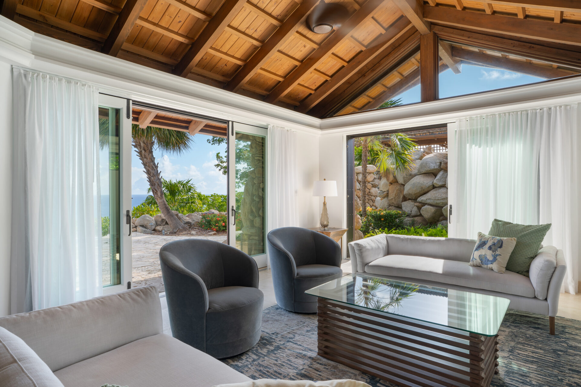 A cozy living room in a villa with a high wooden ceiling and glass doors that open to a garden with palm trees. The room features a light gray sofa, two dark gray armchairs, a glass coffee table, and a floor lamp. Natural light floods the space, enhancing its brightness like wings unfolding.