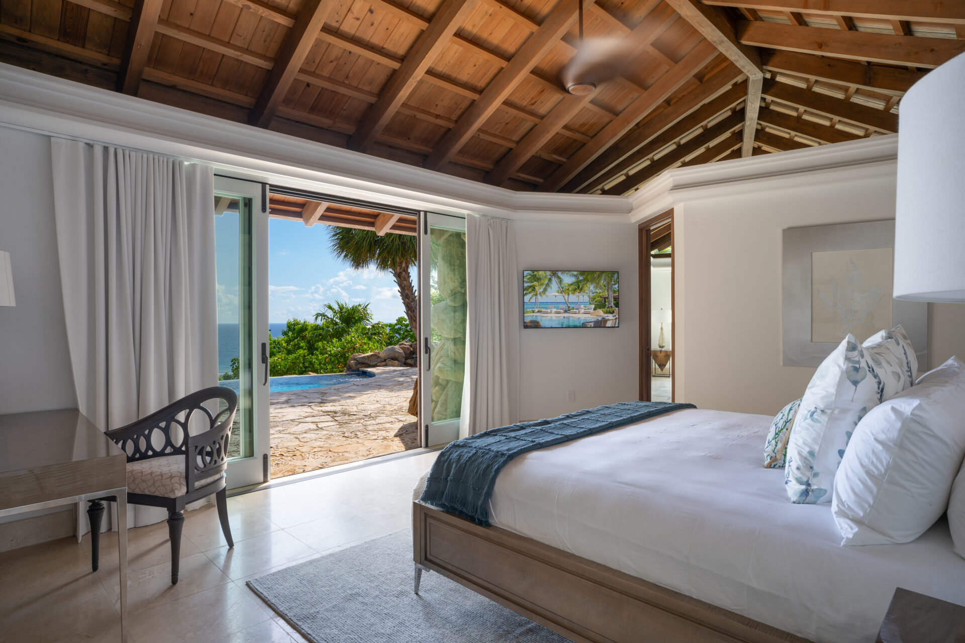 A spacious, airy bedroom at Wings Villa features a wooden ceiling and an open double door leading to a terrace with ocean views. The room has a neatly made bed with blue and white linens, a desk with a chair, and a ceiling fan. The terrace also boasts a pool surrounded by lush greenery.