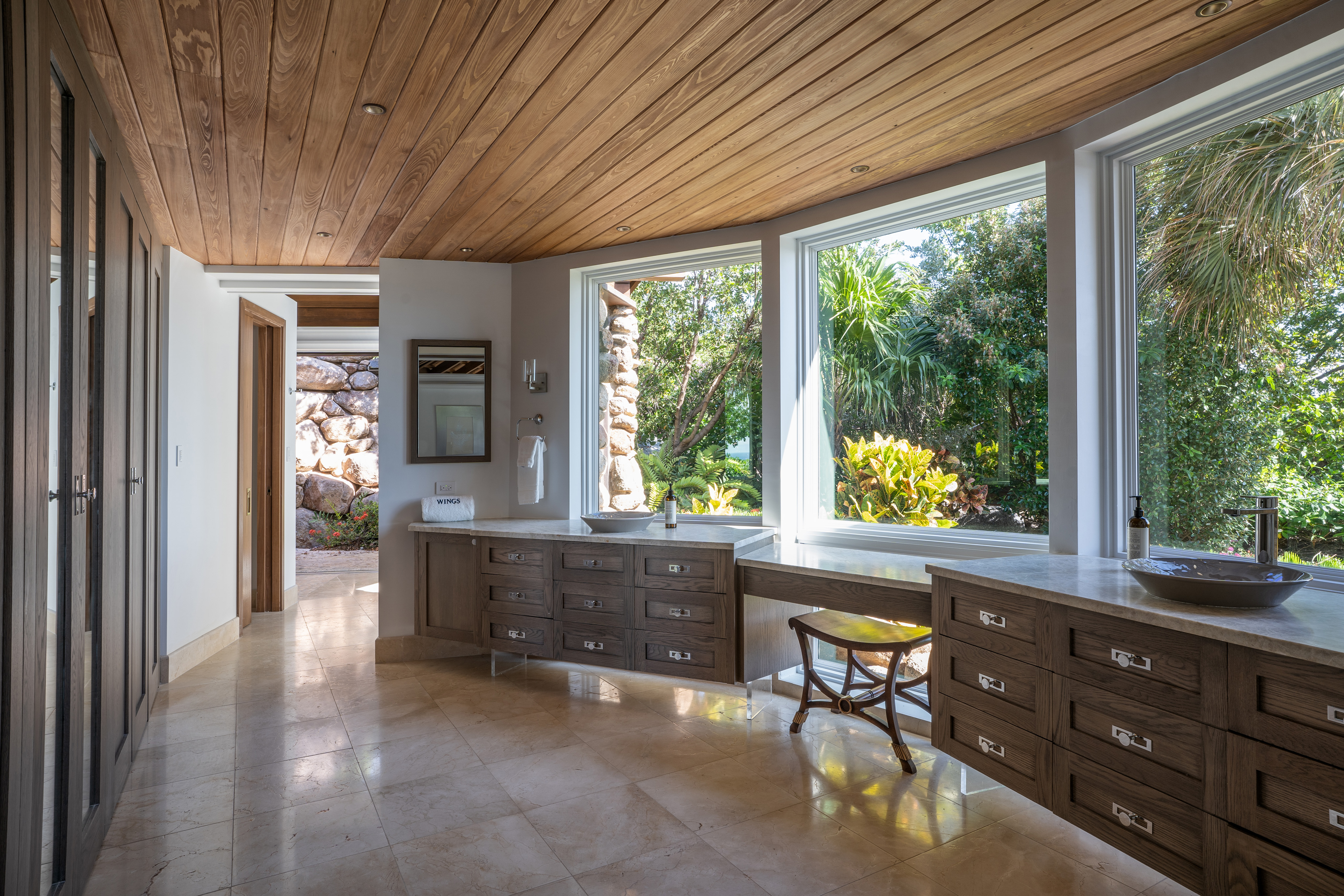 Spacious modern bathroom in a charming villa with large windows showcasing lush outdoor greenery. Features include a wooden ceiling, light tiled flooring, ample counter space with wooden cabinets, and dual sinks. A rustic stool and towel hang by the window, enhancing the serene ambiance.